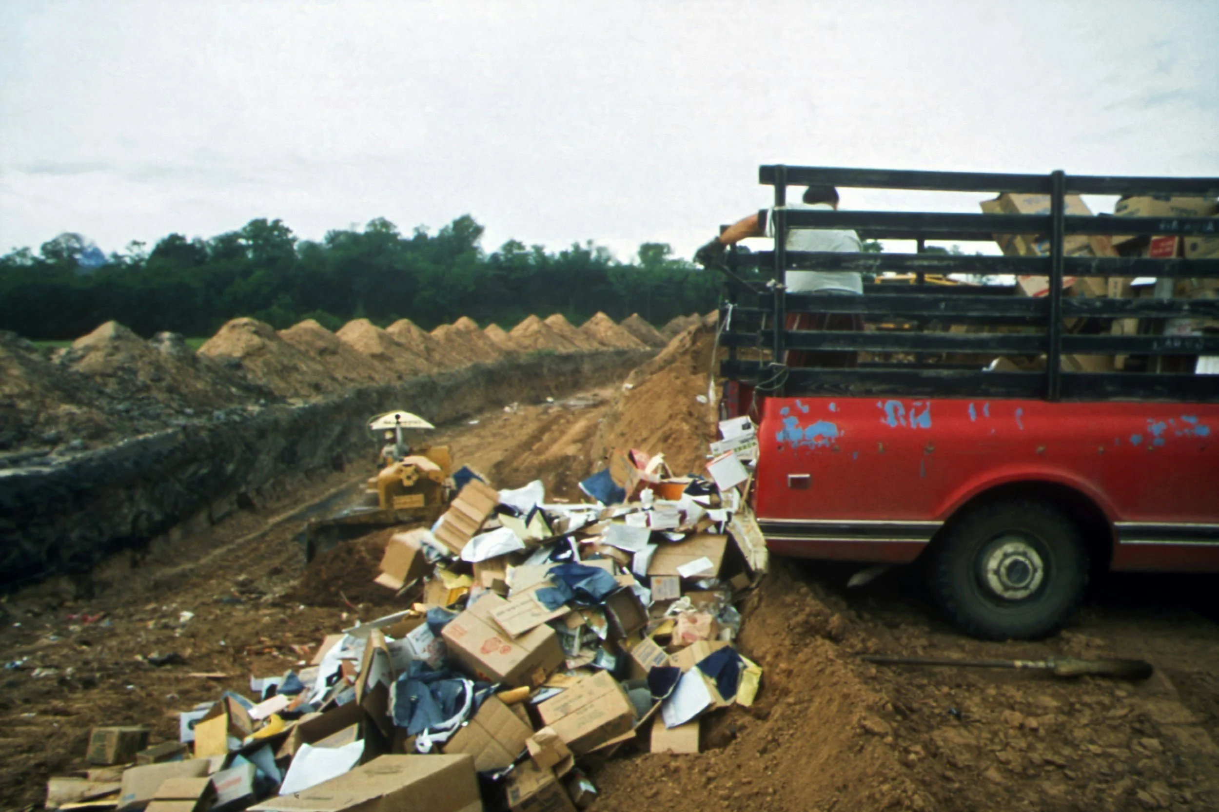 A red truck with black rails is stuck in a muddy construction site, with a pile of discarded cardboard boxes and debris in front of it. In the background, there is a row of dirt mounds and a person standing on the truck, facing away from the camera.