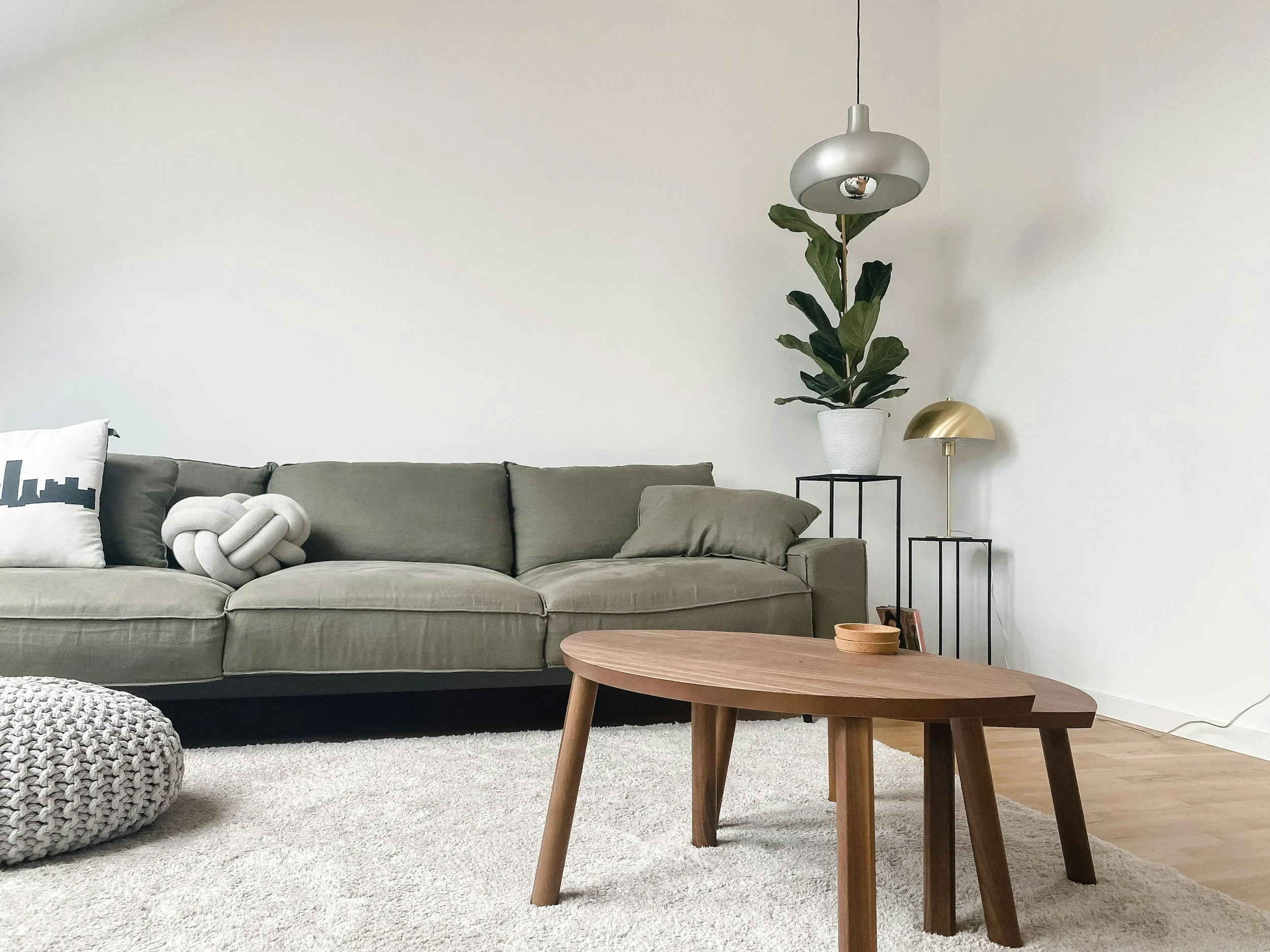 Living room with a light gray sofa, a beige knot pillow, a pillow with a cityscape design, a wooden coffee table, a large potted plant on a black stand, a gold and black modern table lamp, and a gray knitted pouf on a light beige rug. The background features plain white walls.