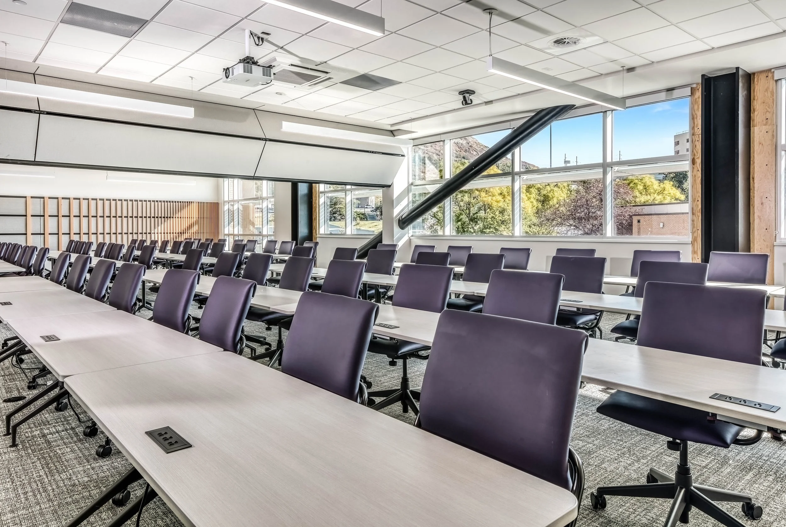 Empty modern conference room with multiple rows of black chairs and white tables, large windows showing trees and buildings outside, and ceiling with lights and a projector.