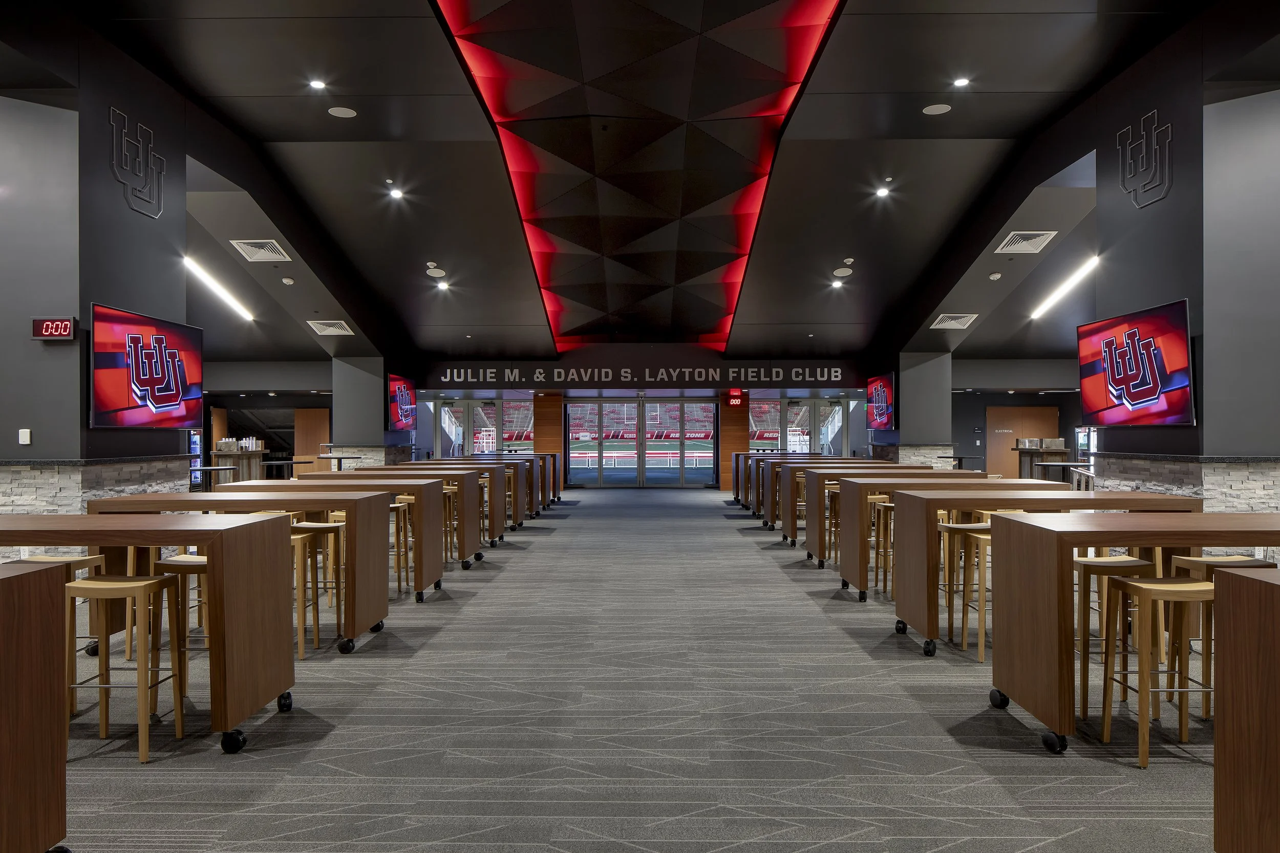 Empty interior of Julie M. & David S. Layton Field Club at a sports stadium, with wooden tables and chairs, multiple TV screens displaying the Indiana University logo, and a view of a sports field through glass doors.
