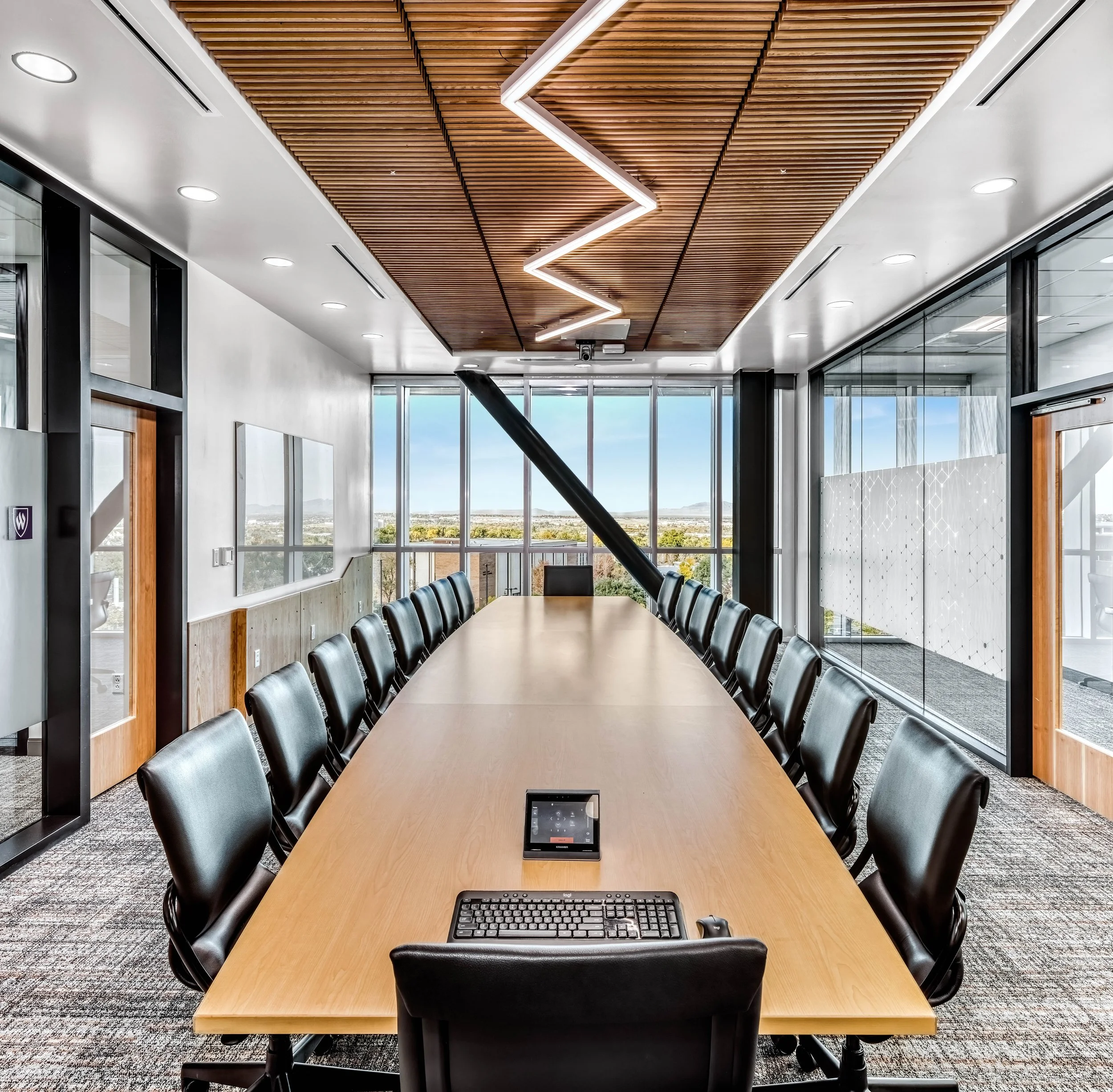 Empty modern conference room with a long wooden table, black office chairs, large windows with a city view, and a decorative ceiling with geometric lighting.