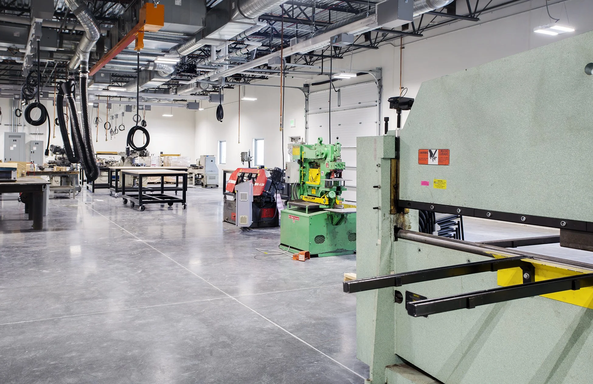 A spacious, well-lit industrial workshop with various manufacturing machines, work tables, and black hoses hanging from the ceiling.