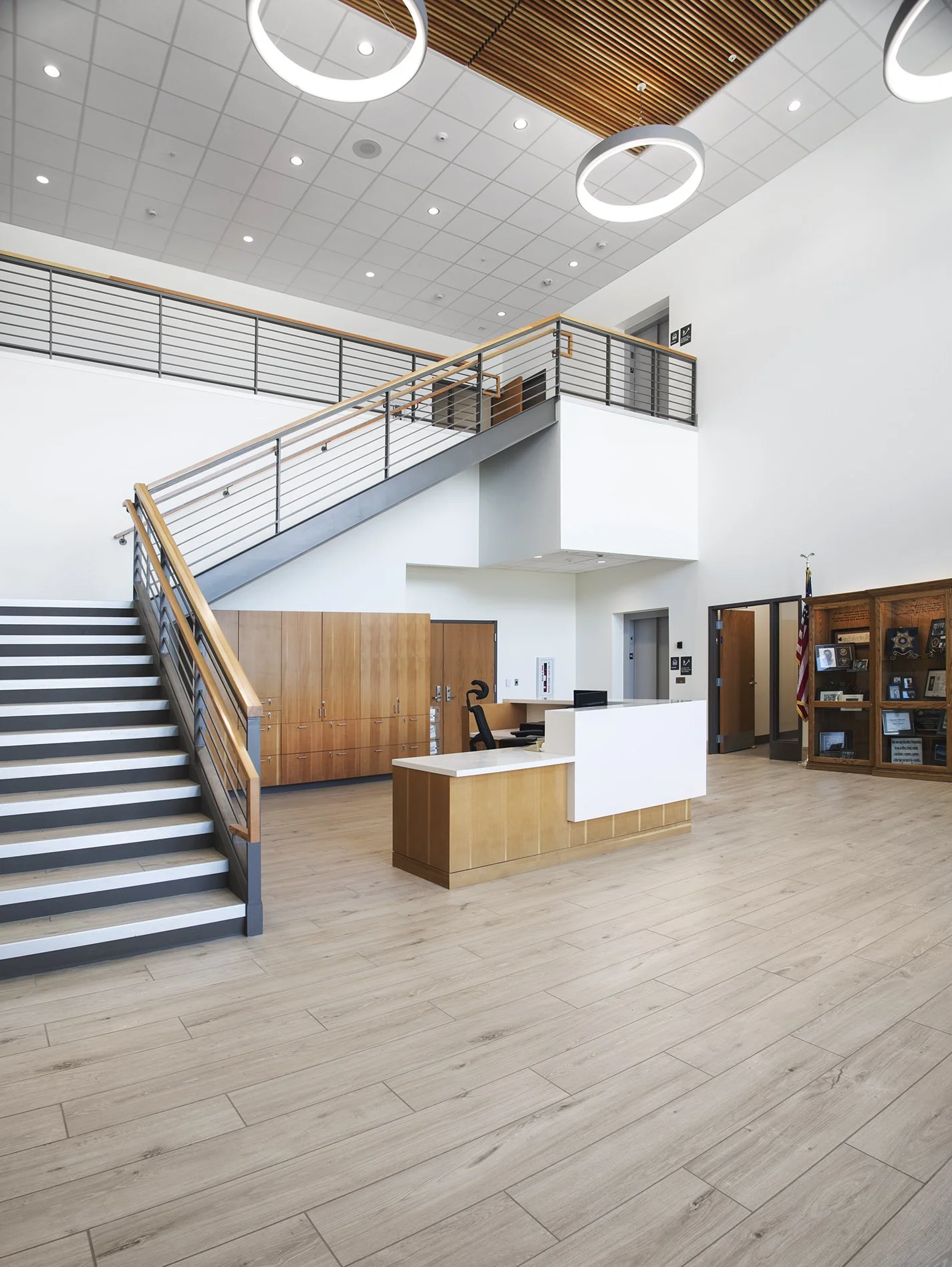 A modern, spacious lobby with a staircase leading to an upper level, a reception desk with an office chair, wooden lockers, and display shelves with framed photographs and certificates.