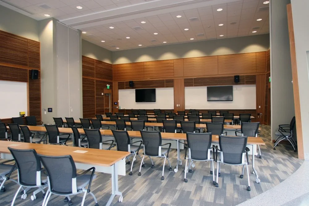 Empty conference room with rows of black chairs, wooden tables, two large TV screens on the wall, and wood-paneled walls.