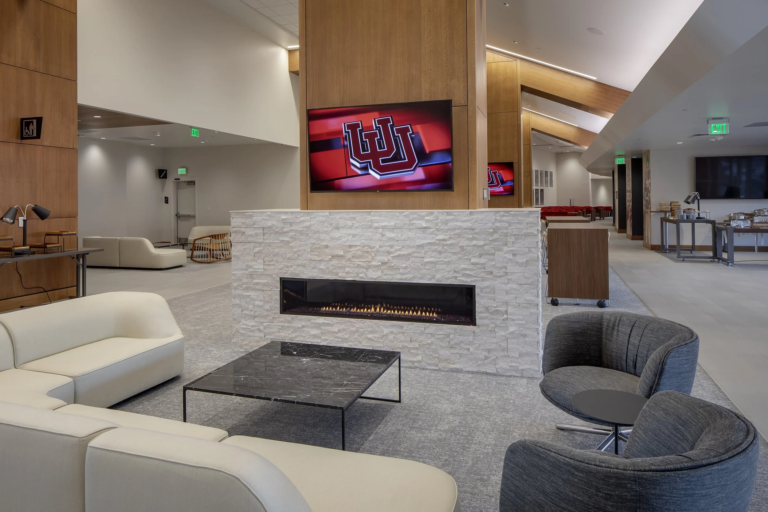 Modern hotel lobby with a white stone fireplace, a flat-screen TV displaying the University of Utah logo, and comfortable seating including a white sofa and gray chairs.