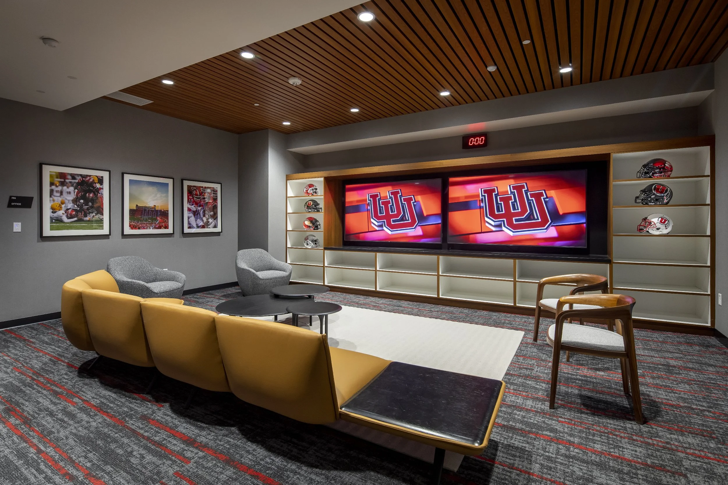 A modern lounge room with a large screen TV displaying the Utah Utes logo, football helmets on shelves, yellow and gray chairs, coffee tables, and framed sports photos on the wall.