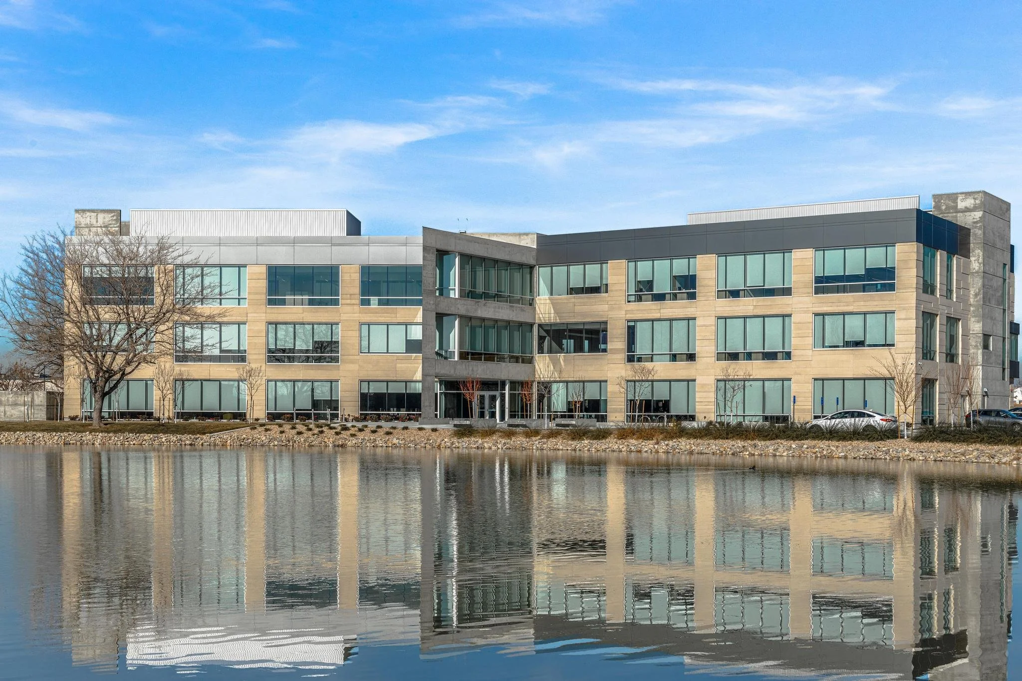 Modern office building with large glass windows reflected in a body of water in the foreground, with a blue sky and some trees around.