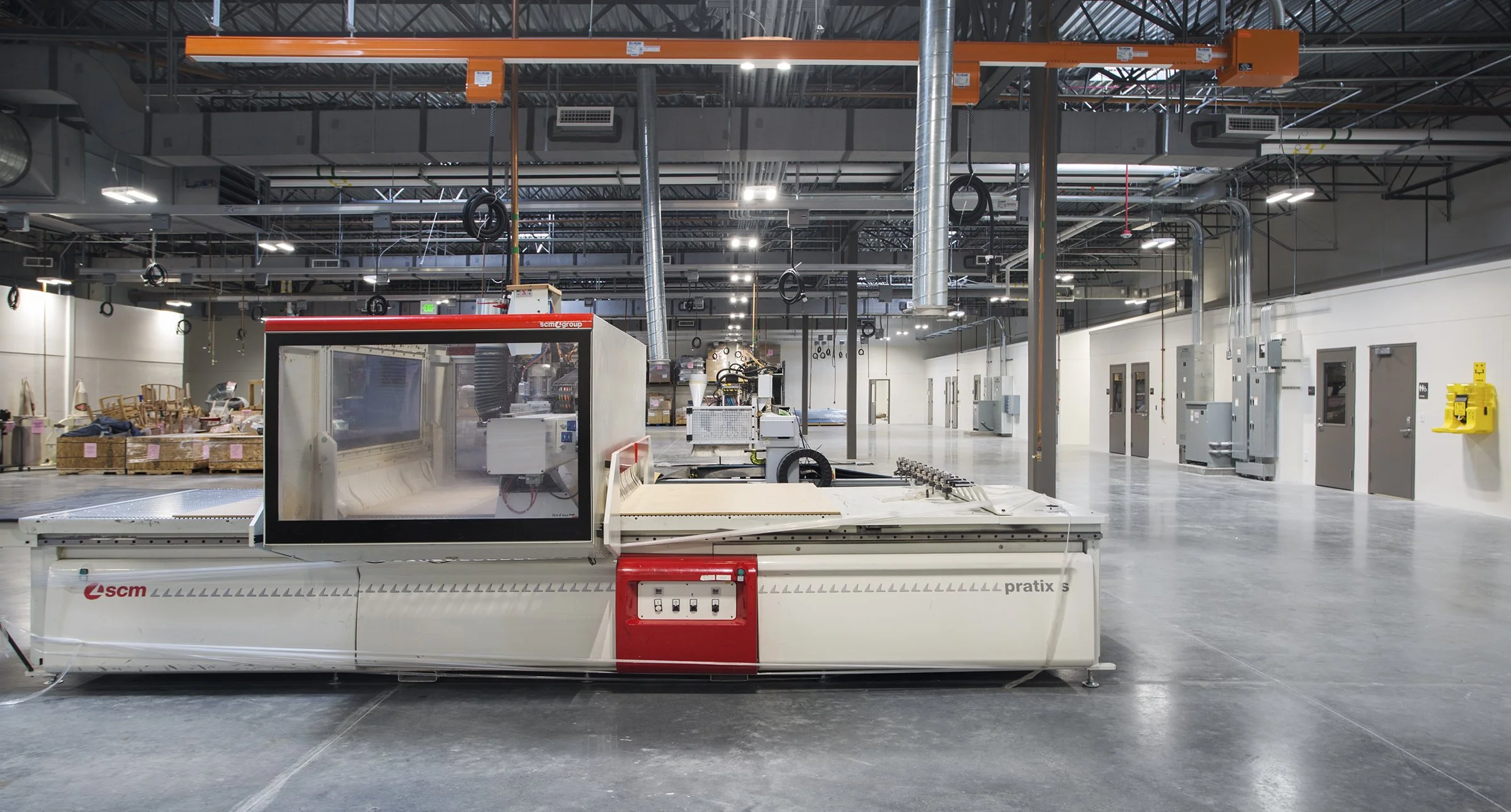 Empty industrial warehouse with large manufacturing machine in the foreground, several closed doors along the back wall, and various industrial equipment and boxes in the background, with high ceiling and exposed ductwork.