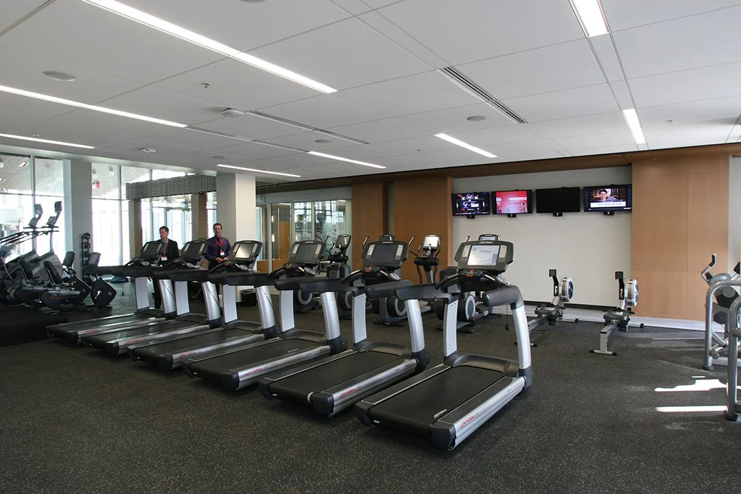 Indoor gym with multiple treadmills and rowing machines facing a wall with three TV screens, windows on the left, and two staff members in the background.