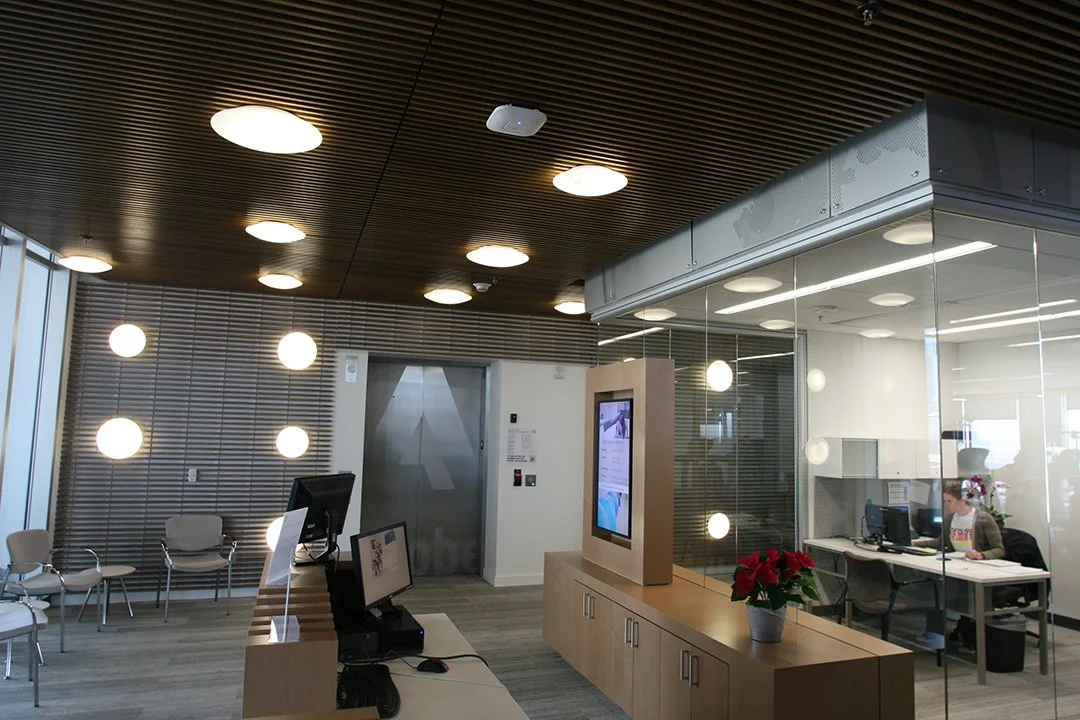 Modern office lobby with waiting chairs, a reception desk, a glass-enclosed office with a person working at a desk, and decorative lighting on the ceiling and wall.