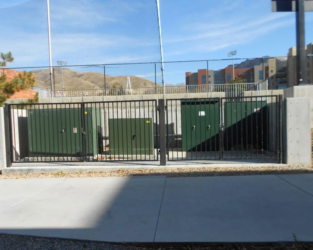 A chain-link fence gate with large green electrical boxes inside, set against a concrete wall with a sports field and mountains in the background.