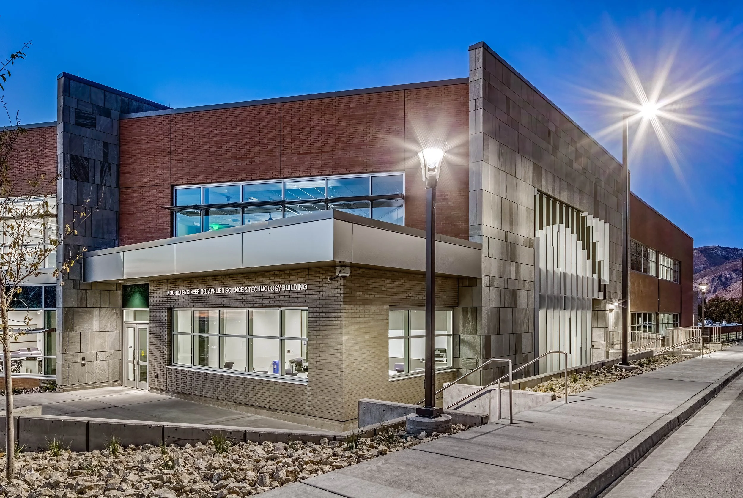 Exterior of the Noordra Engineering, Applied Science & Technology Building at dusk, with street lamps illuminated and mountains in the background.