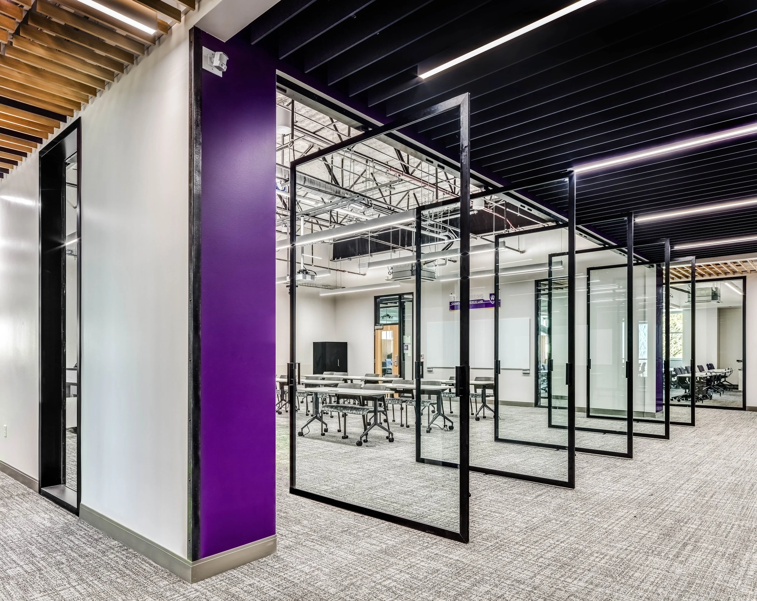 Modern office space with glass-walled conference rooms, purple accent wall, black ceiling slats with integrated lighting, and carpeted flooring.