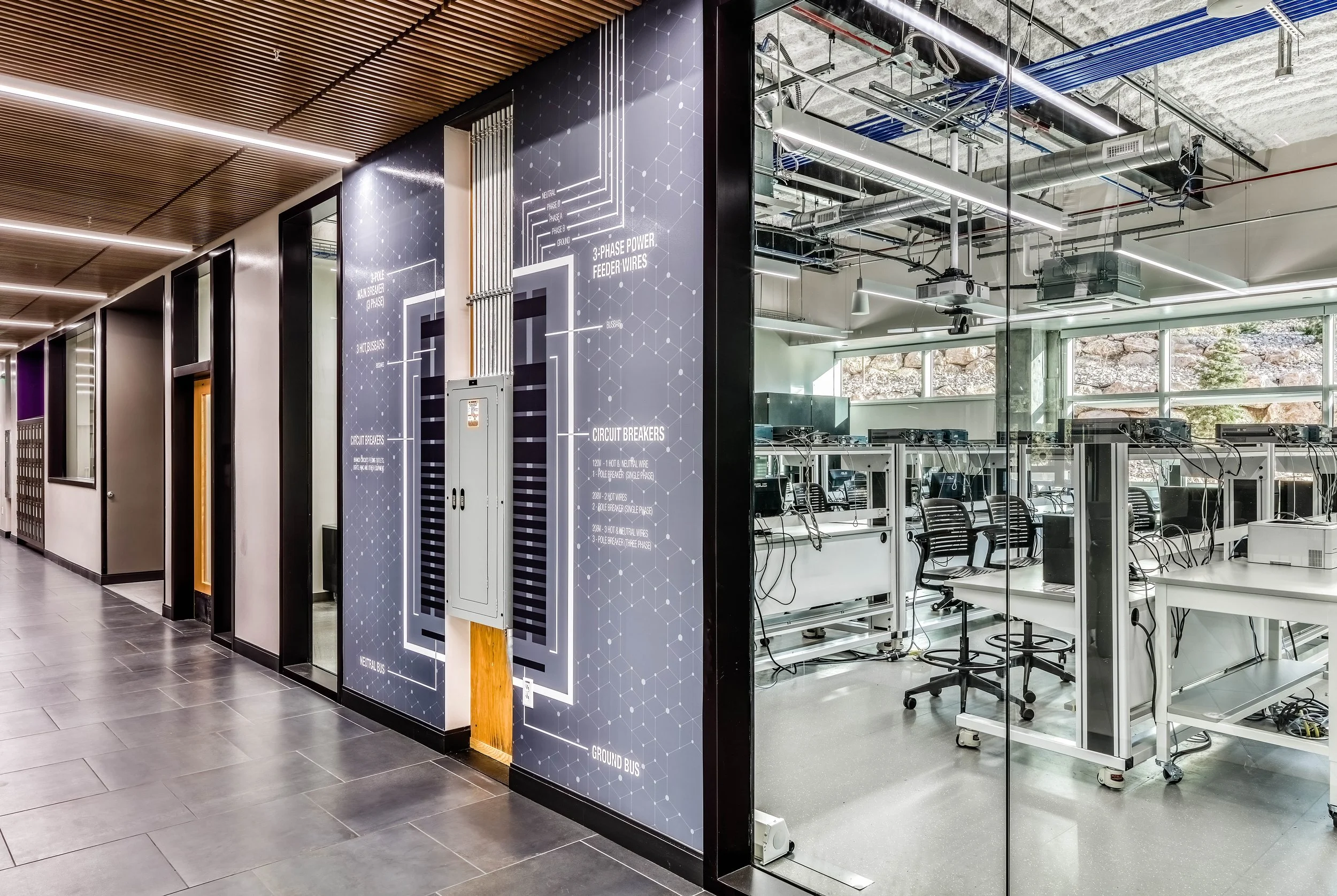 Modern office hallway with a glass enclosure showing multiple workstations with computers and chairs, and technical wiring on the ceiling. The hallway features wood-paneled ceiling and gray tile flooring, with a poster displaying electrical circuit d