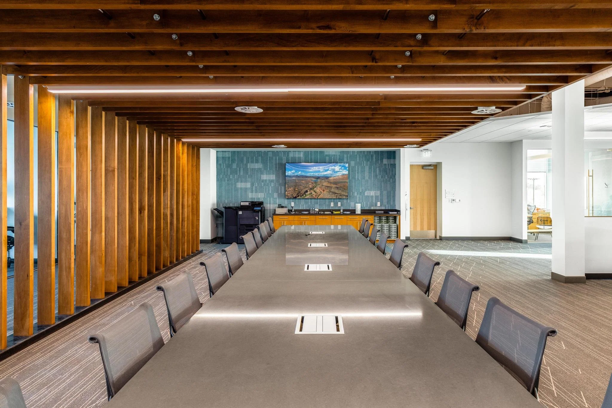 A modern conference room with a long rectangular table, multiple chairs, a wall-mounted screen displaying a landscape, and a wooden slatted ceiling.