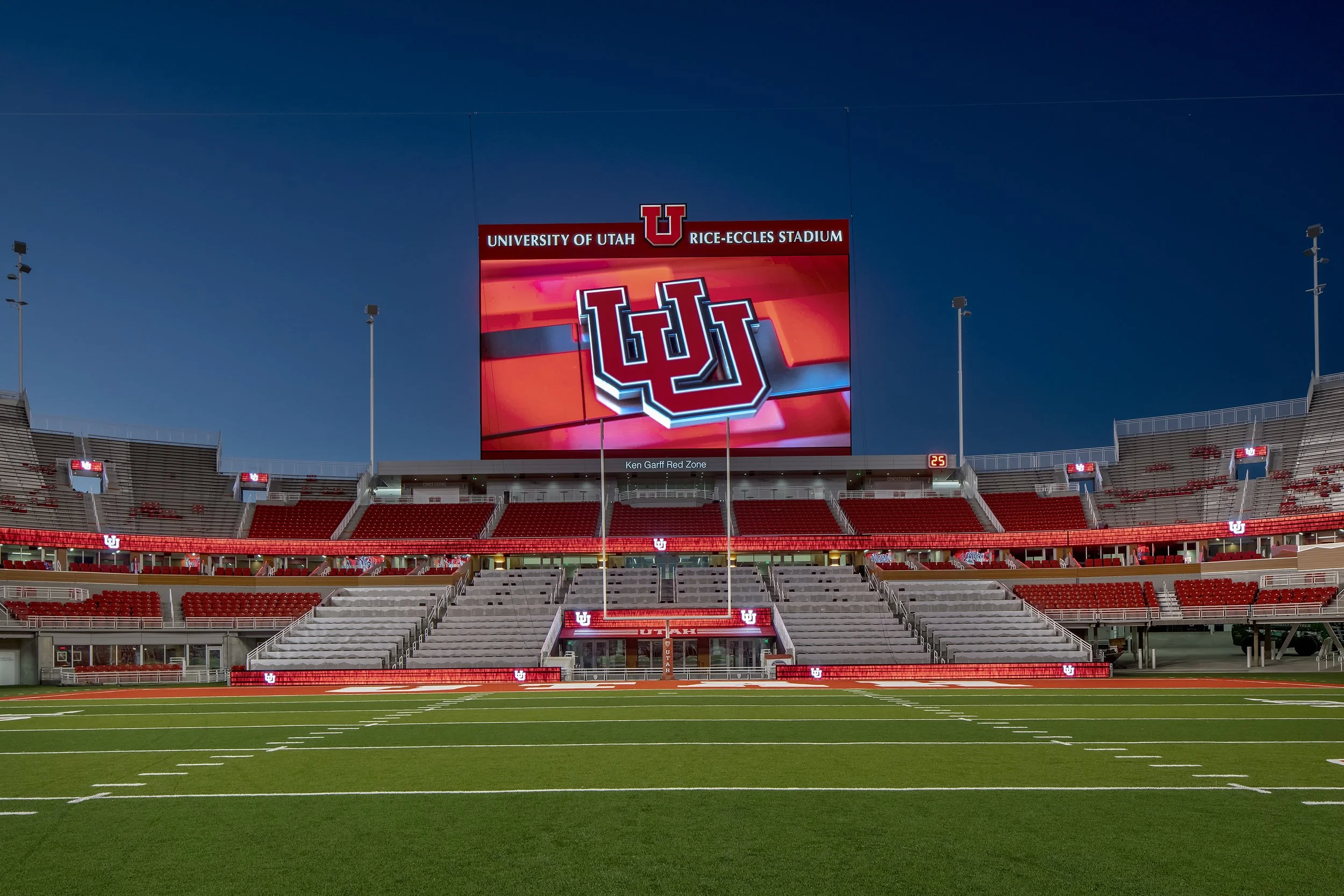 Empty football stadium with a large digital screen displaying the University of Utah logo, red and white colors, and the words 'University of Utah Rice-Eccles Stadium' at the top, during sunset or twilight.