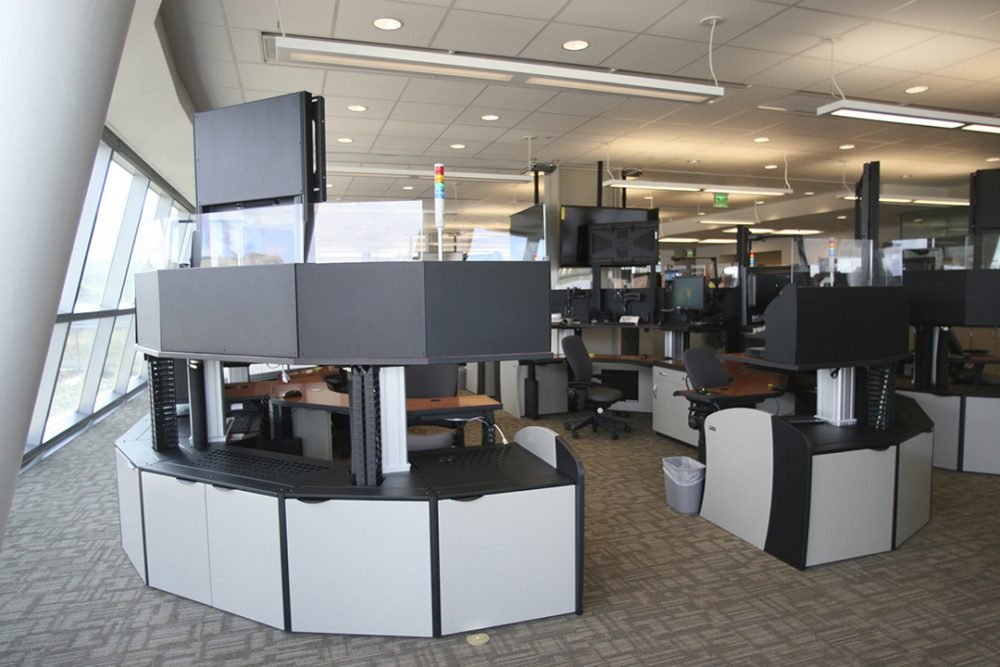 Airport security checkpoint with multiple screening stations, monitors, and chairs in a spacious area with large windows.