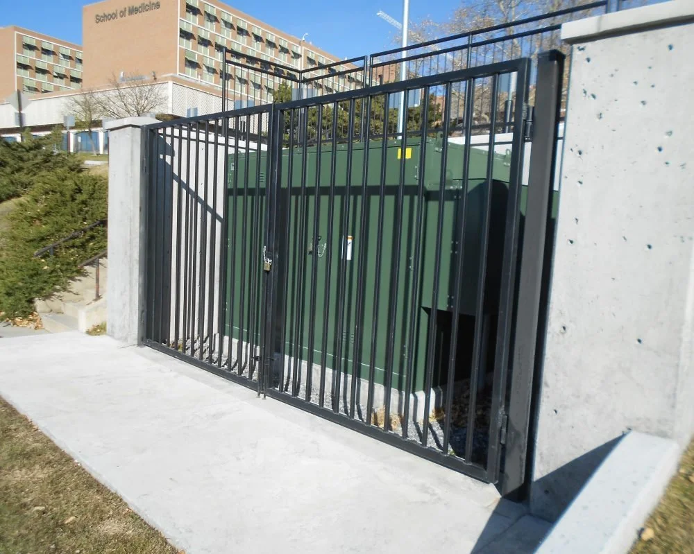 A black metal gate with bars, secured around a green utility box or electrical transformer, located outside near a concrete sidewalk. In the background, there is a large building with a sign that reads 'School of Medicine,' and some trees and a clear