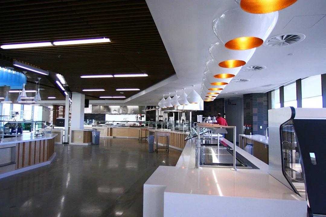 Empty cafeteria with modern lighting and design, including white and orange hanging lamps, a serving counter, and a person in red working in the background.