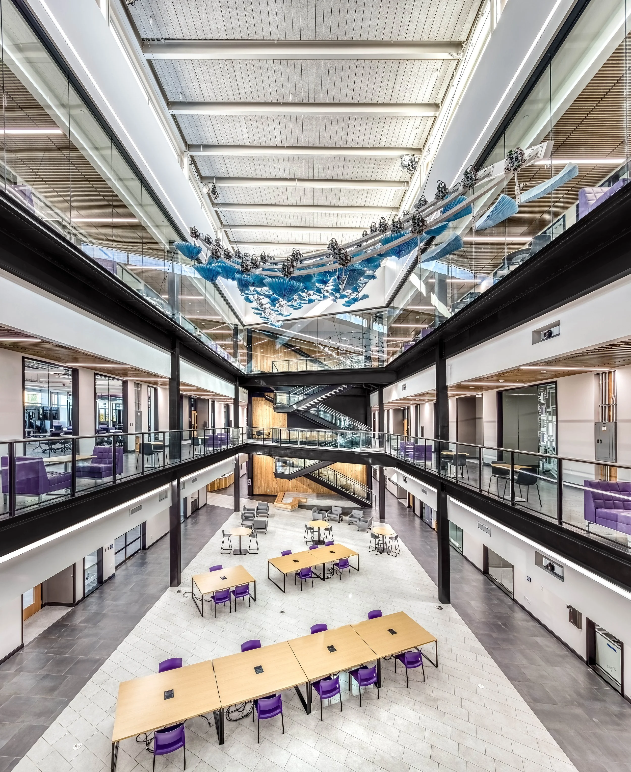 Interior view of a modern, multi-story office building with an open atrium, glass railings, purple chairs, wooden tables, and hanging blue art installation resembling a bird in flight.