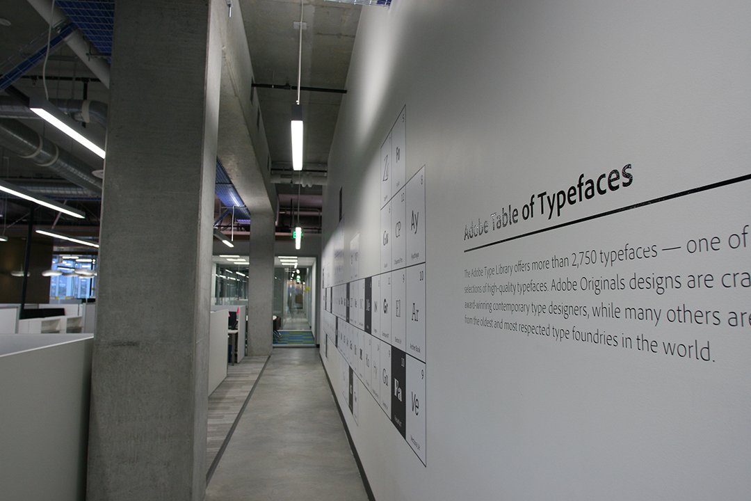 Indoor hallway with a white wall displaying the Adobe Table of Typefaces, and text describing Adobe's font library, in a modern office setting with concrete and glass design elements.