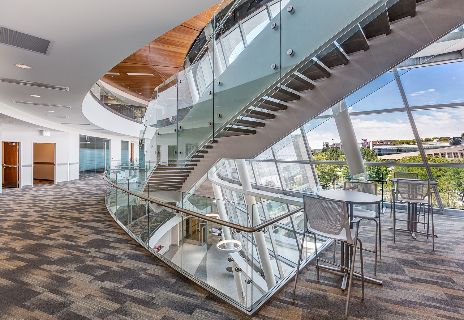 Modern office building interior with curved glass staircase, large floor-to-ceiling windows, and a small seating area with tables and chairs.