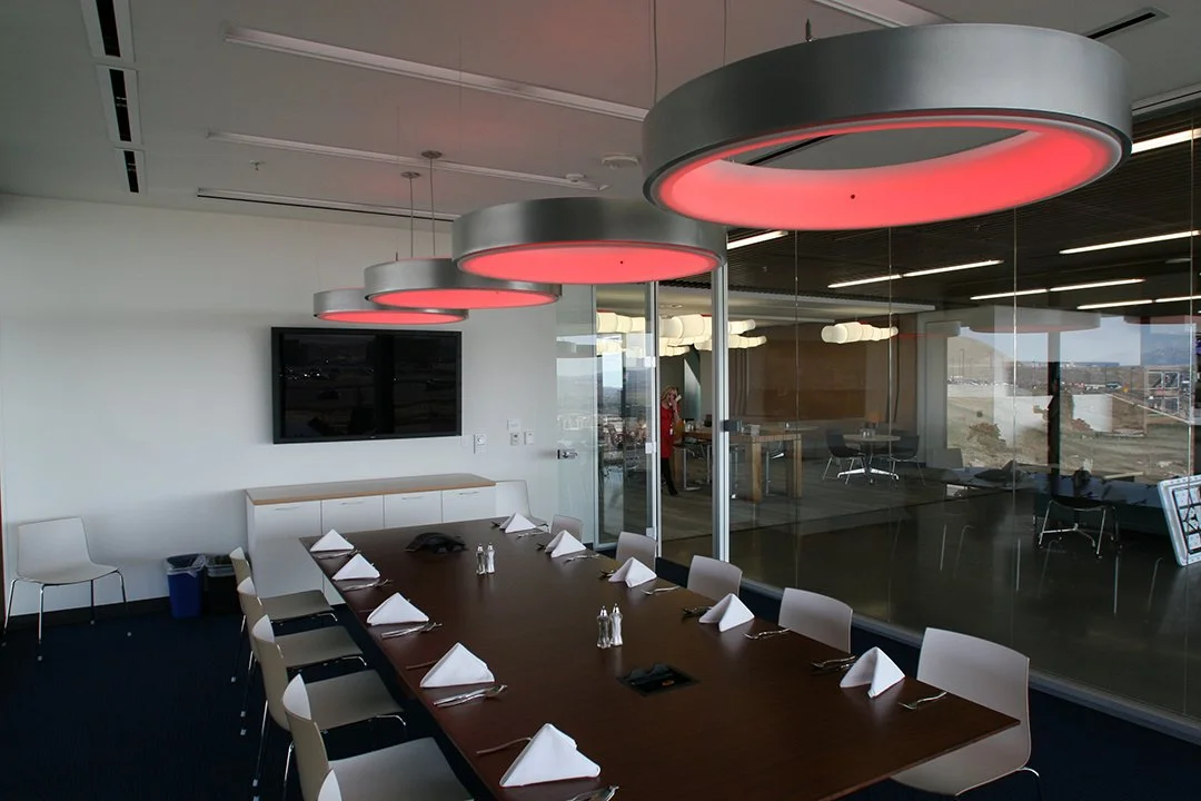 Modern conference room with a large wooden table set for a meeting, white chairs, a wall-mounted TV, and ceiling lights with red rings.
