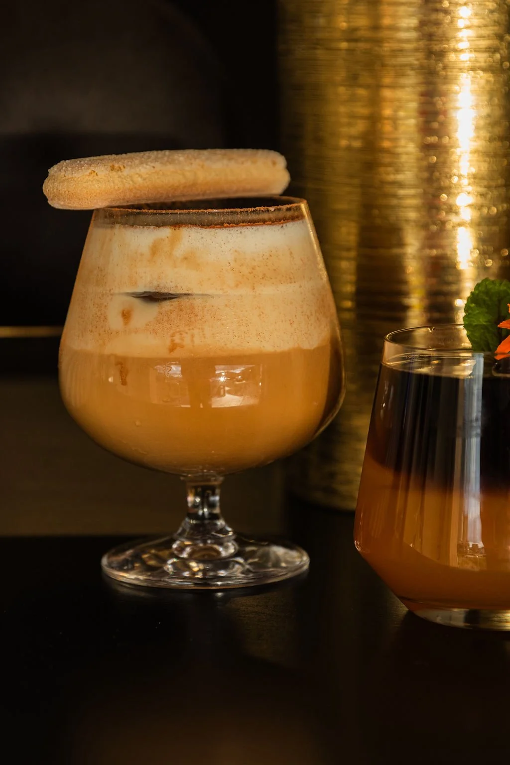 A dessert drink topped with a cookie in a round glass, with a second drink in a taller glass nearby, set against a dark background with warm lighting.