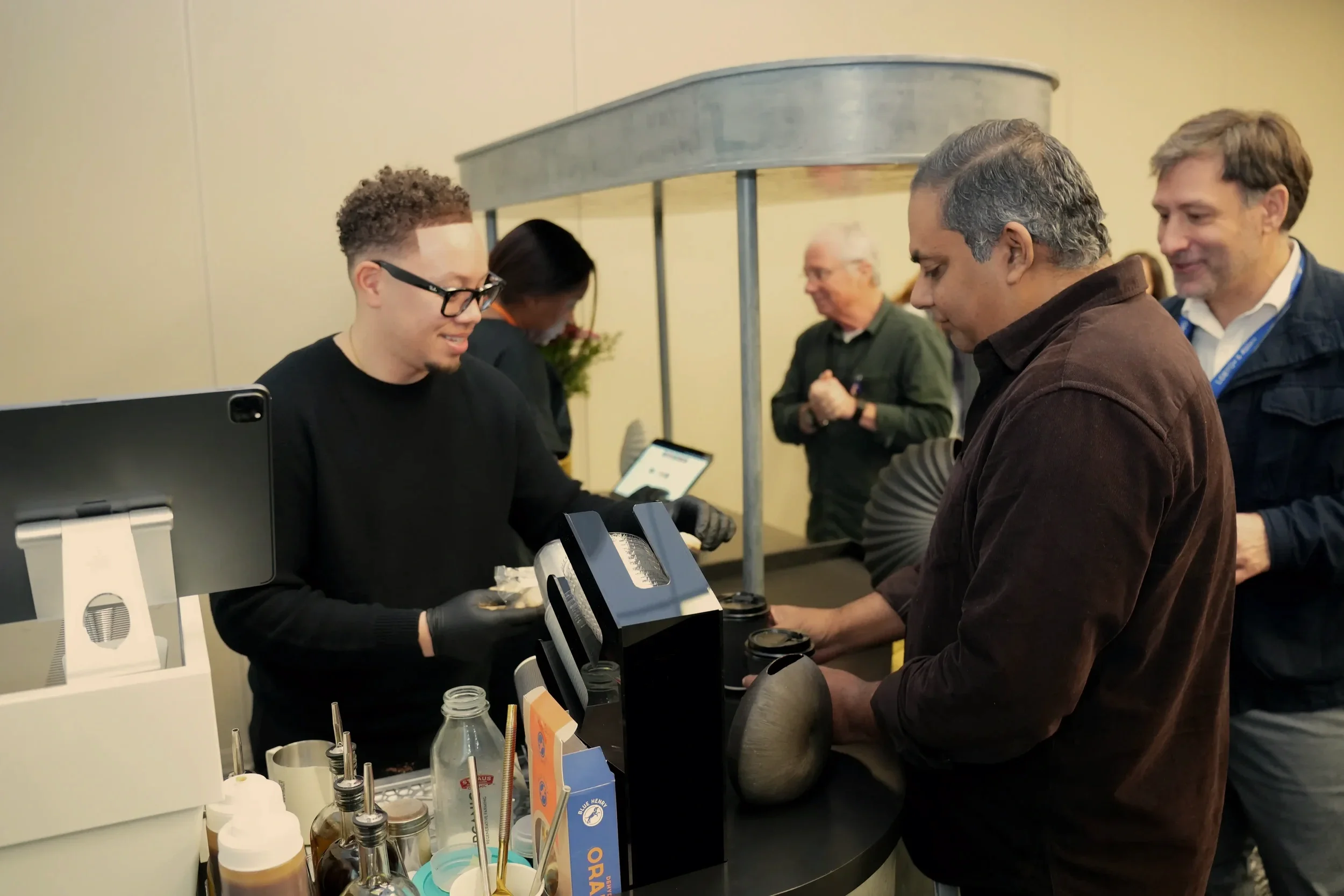 A man at a coffee stand hands a drink to a customer while others wait in line behind them.