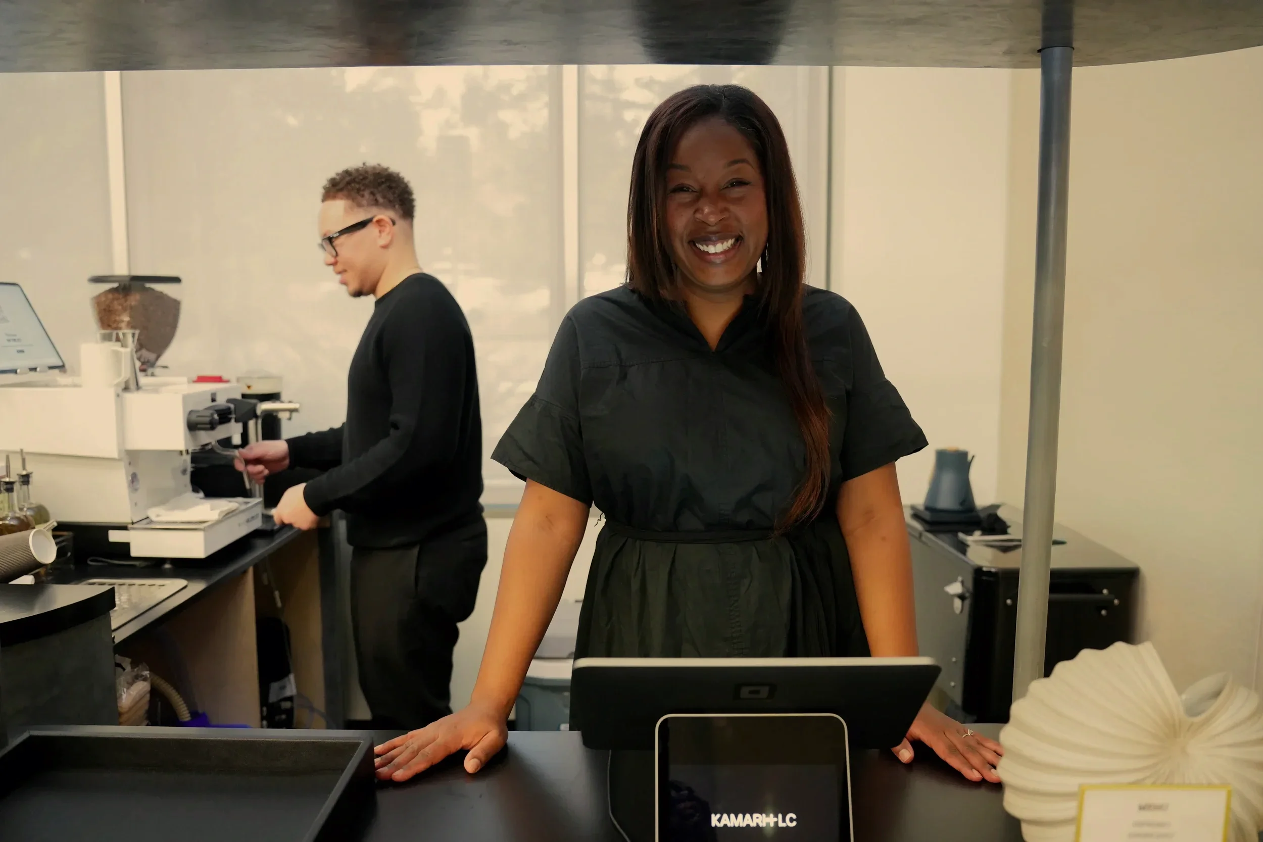 A woman smiling behind a counter with a tablet, with a man working with coffee equipment in the background.