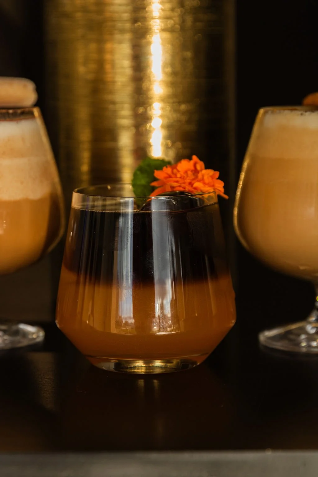 Close-up of three glasses with drinks, one with a dark beverage, two with light, frothy drinks on a black surface, with a blurred metallic background and a small orange flower and green leaf in the center.