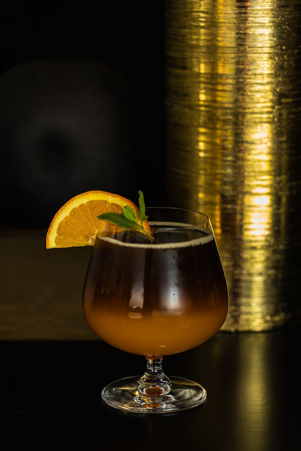 A glass of dark cocktail garnished with an orange slice and mint leaves, set against a dark background with gold decorative elements.