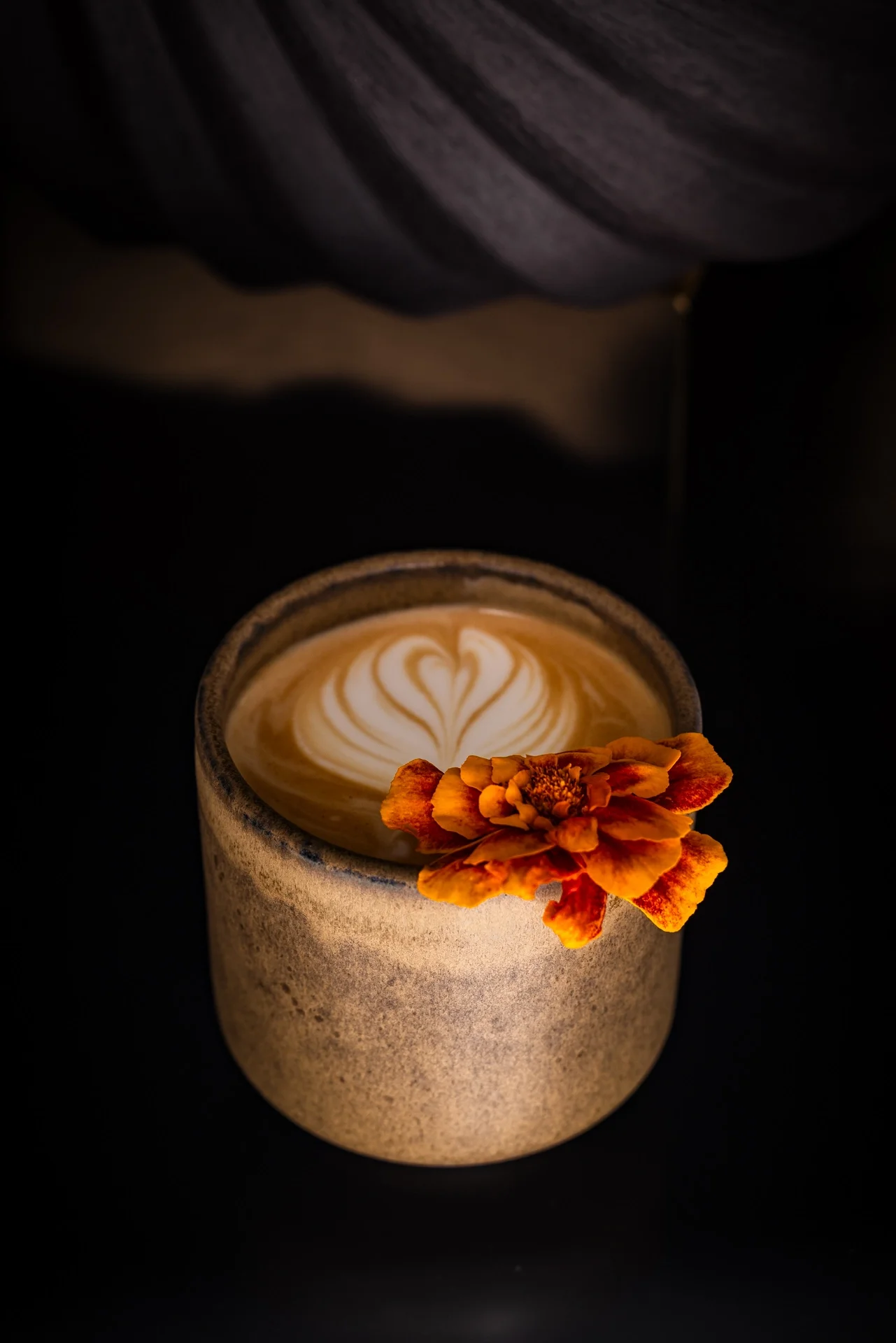 A cup of coffee with latte art and a marigold flower on the rim, placed on a dark surface.