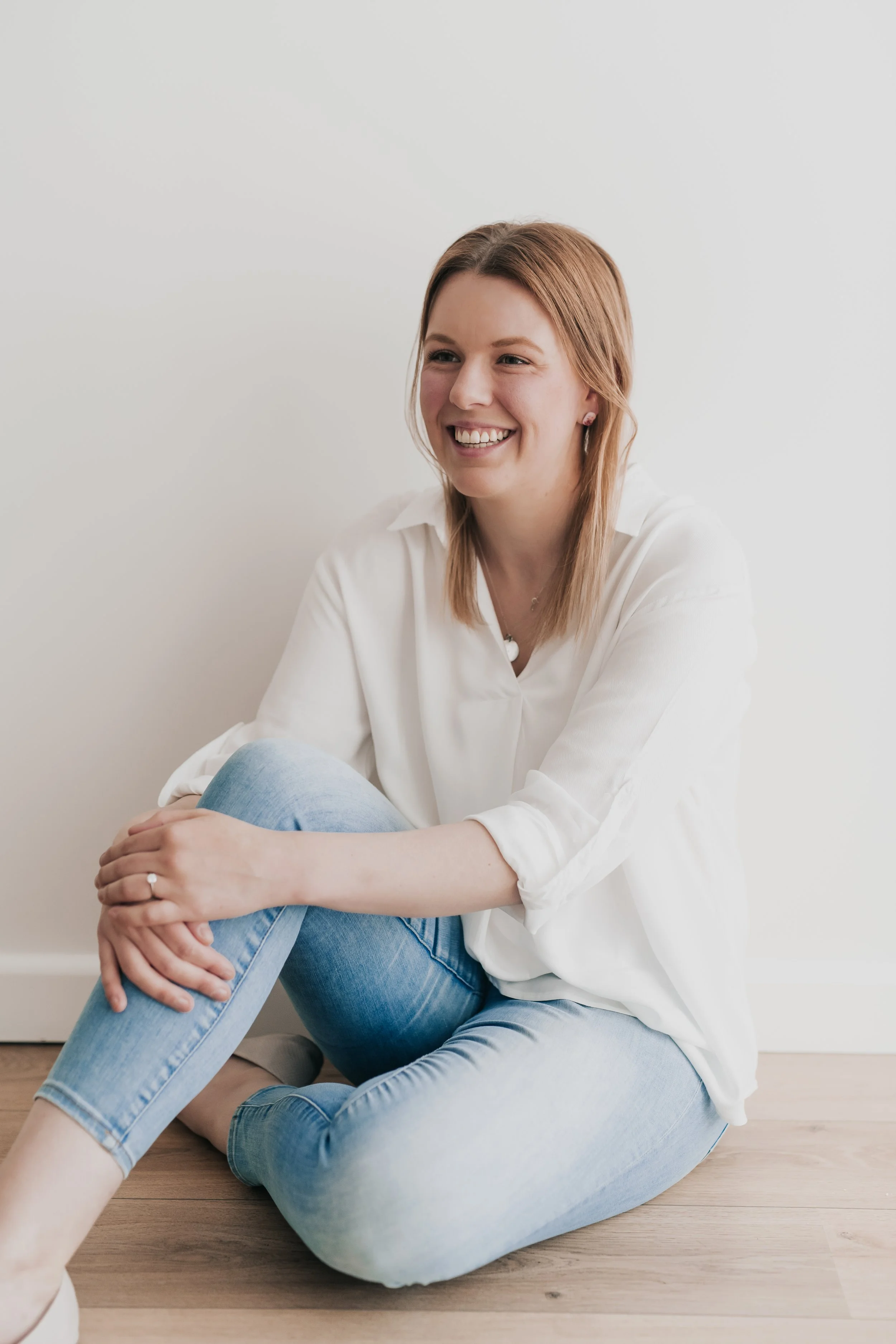 A woman with red hair sitting on the floor, smiling, wearing a white blouse and light blue jeans.