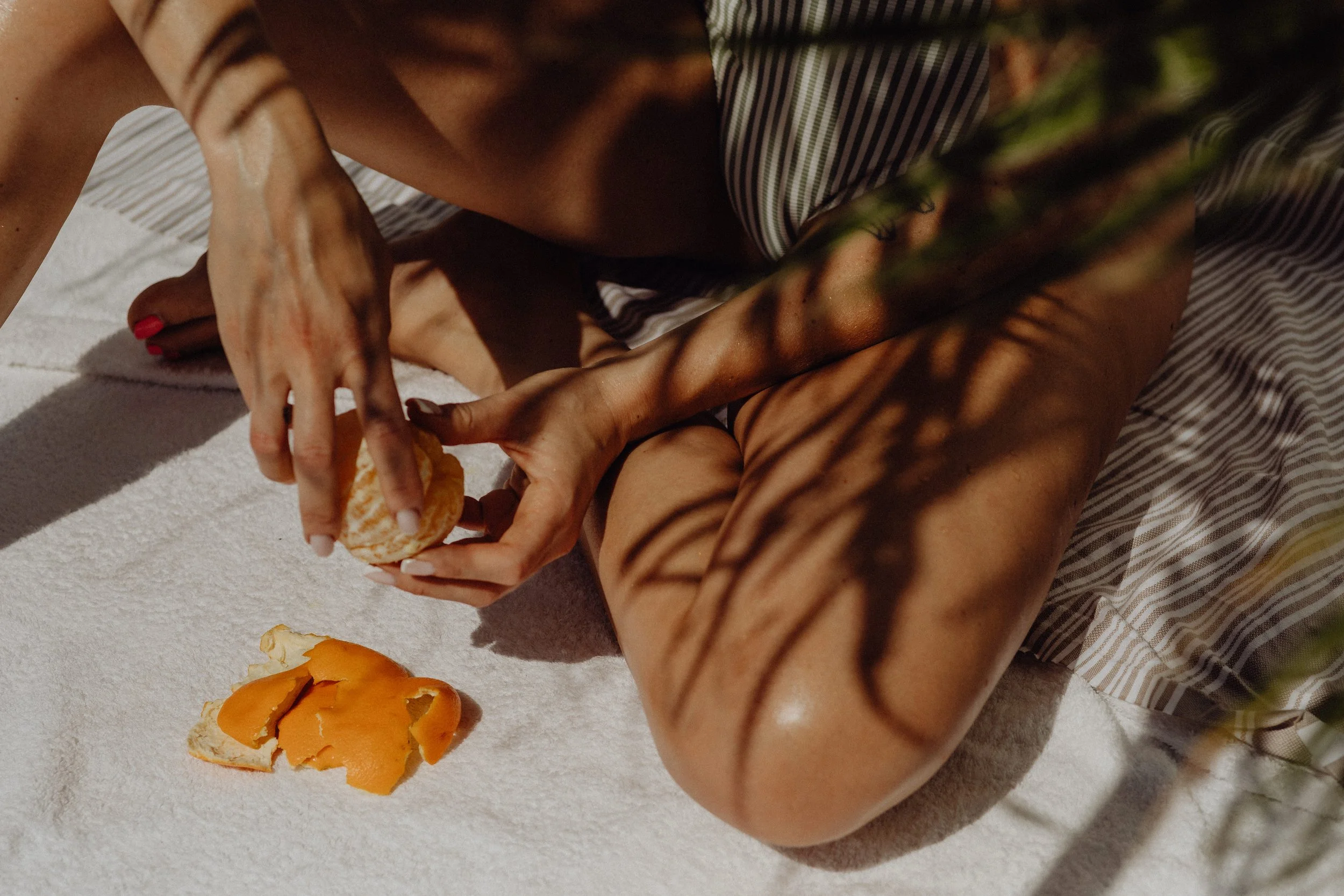 Person peeling oranges on a white towel, with orange peel on towel and striped cloth nearby, shadows of leaves cast on person and towel.