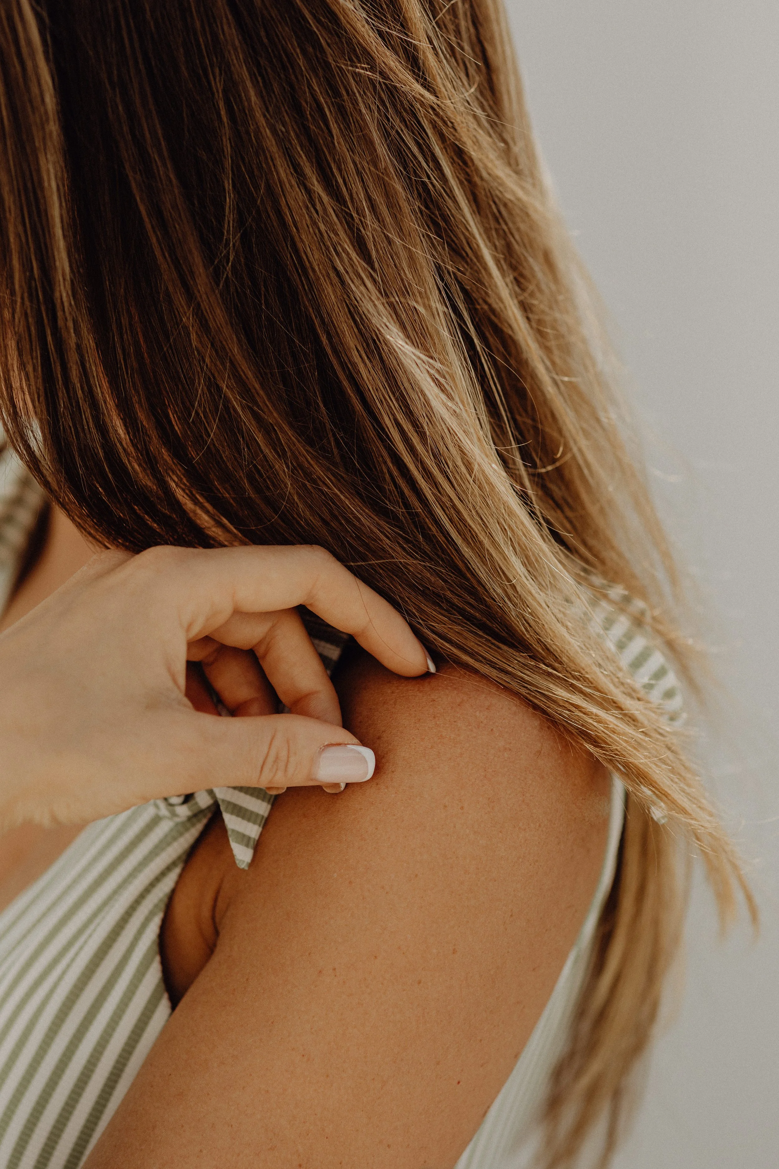 Close-up of a woman with reddish-brown hair touching her shoulder with her right hand, wearing a sleeveless top with grey and white stripes.