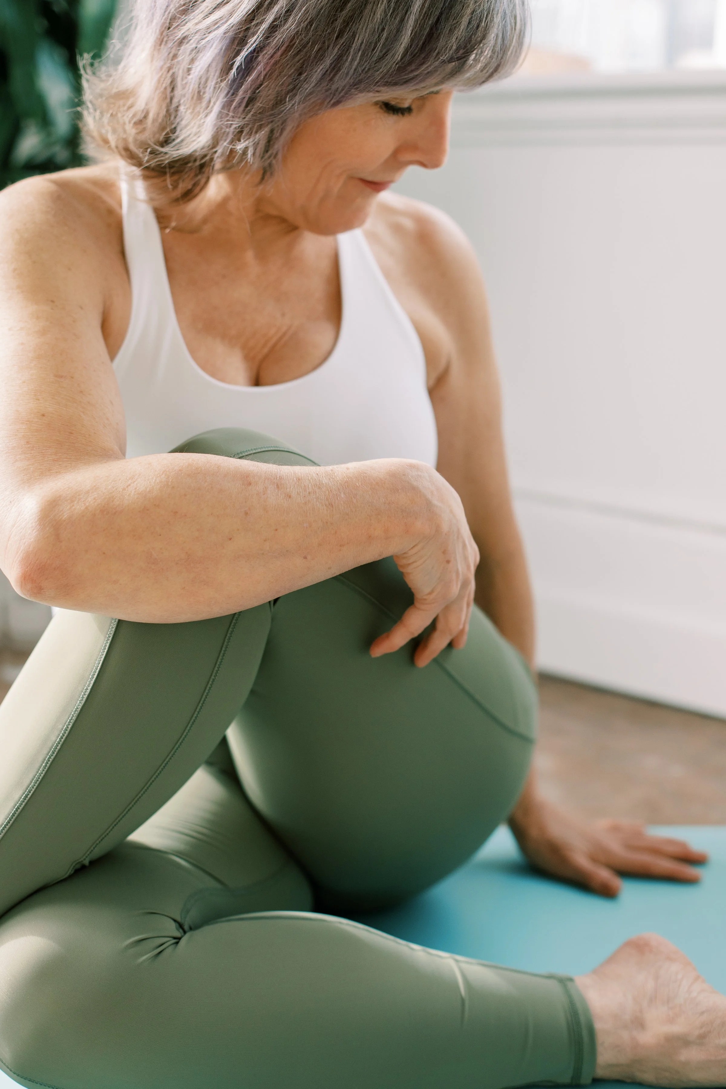 Older woman practicing yoga, sitting on a yoga mat with a green exercise ball, in a bright, airy room.