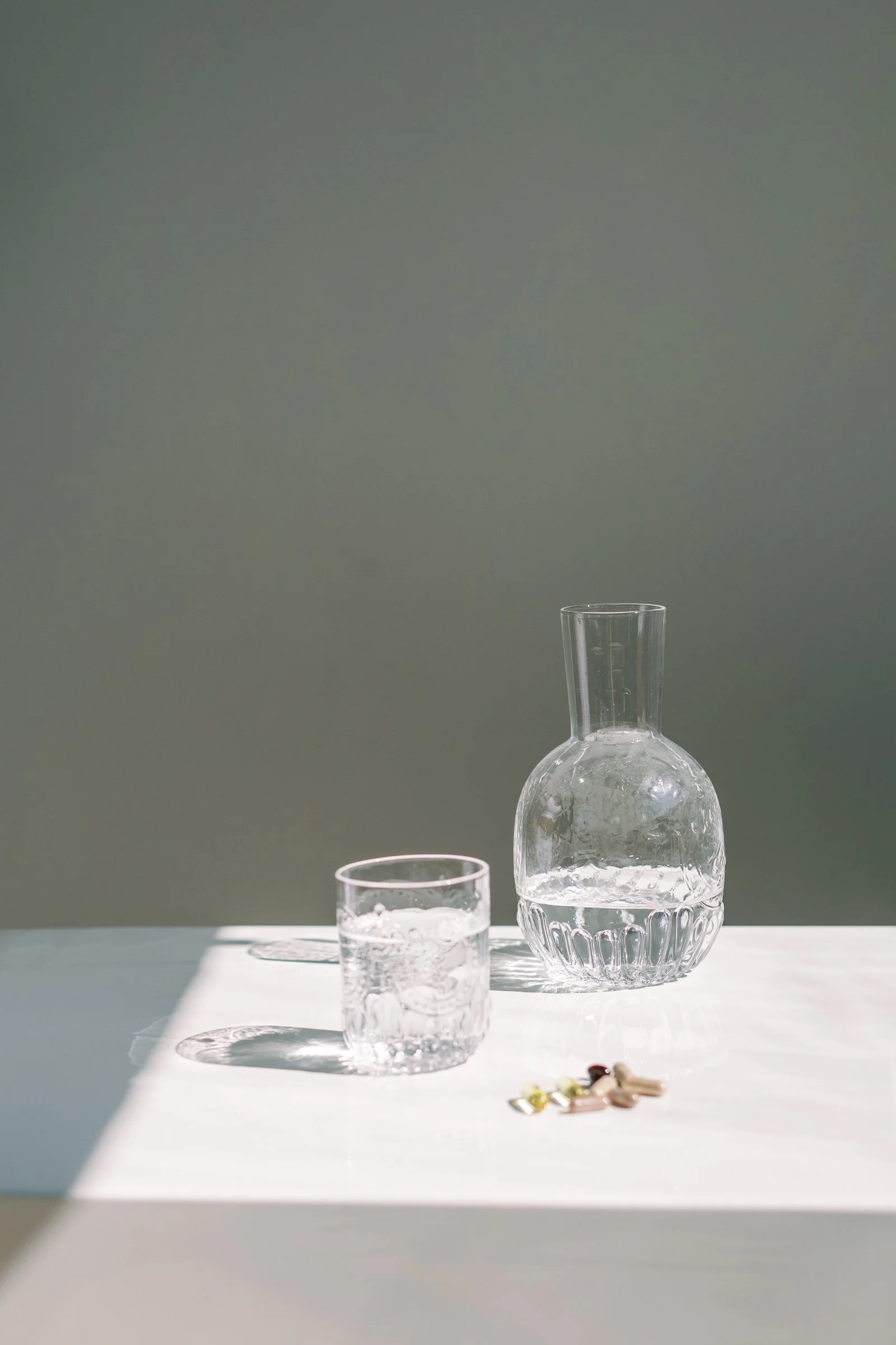 A clear glass water pitcher and a glass of water on a white surface with pills beside them, and shadow and sunlight creating a simple minimalist scene.