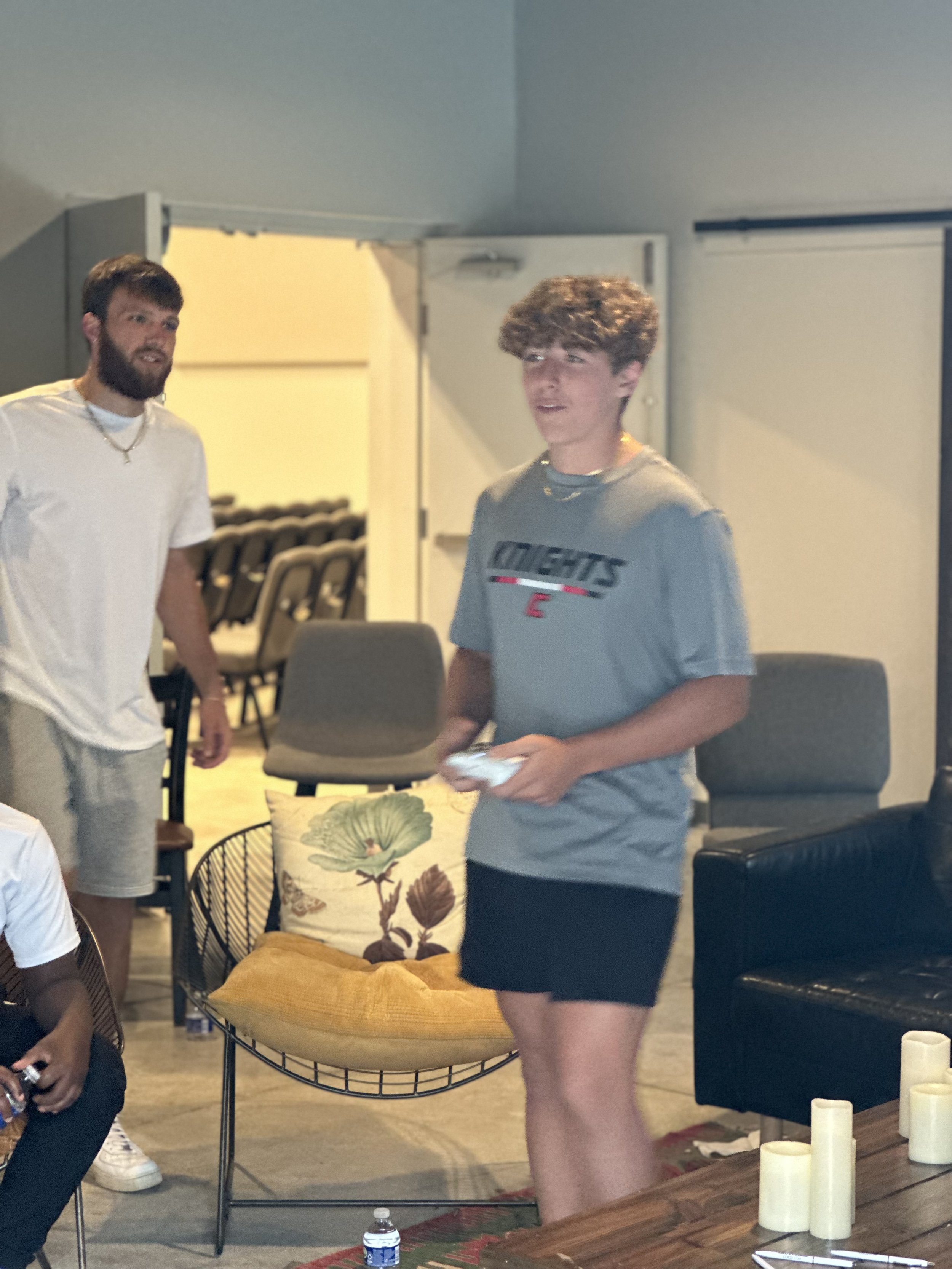 Two young men in casual clothing standing indoors, one holding a game controller. The background features chairs and a decorative pillow on a chair.