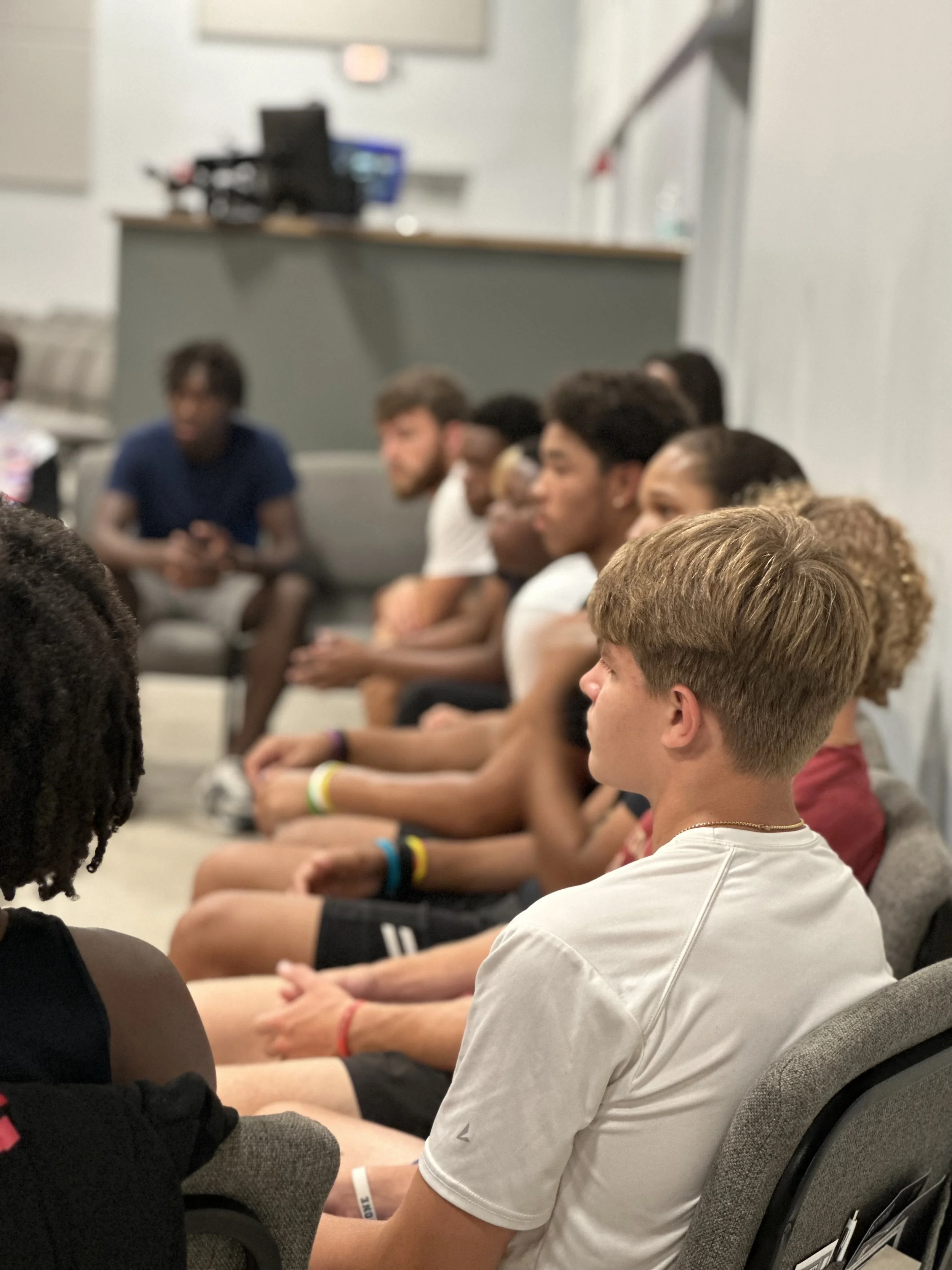 A group of young people sitting in a room, attentively listening to someone or something out of view, with blurred background showing a wall and a raised platform or stage.