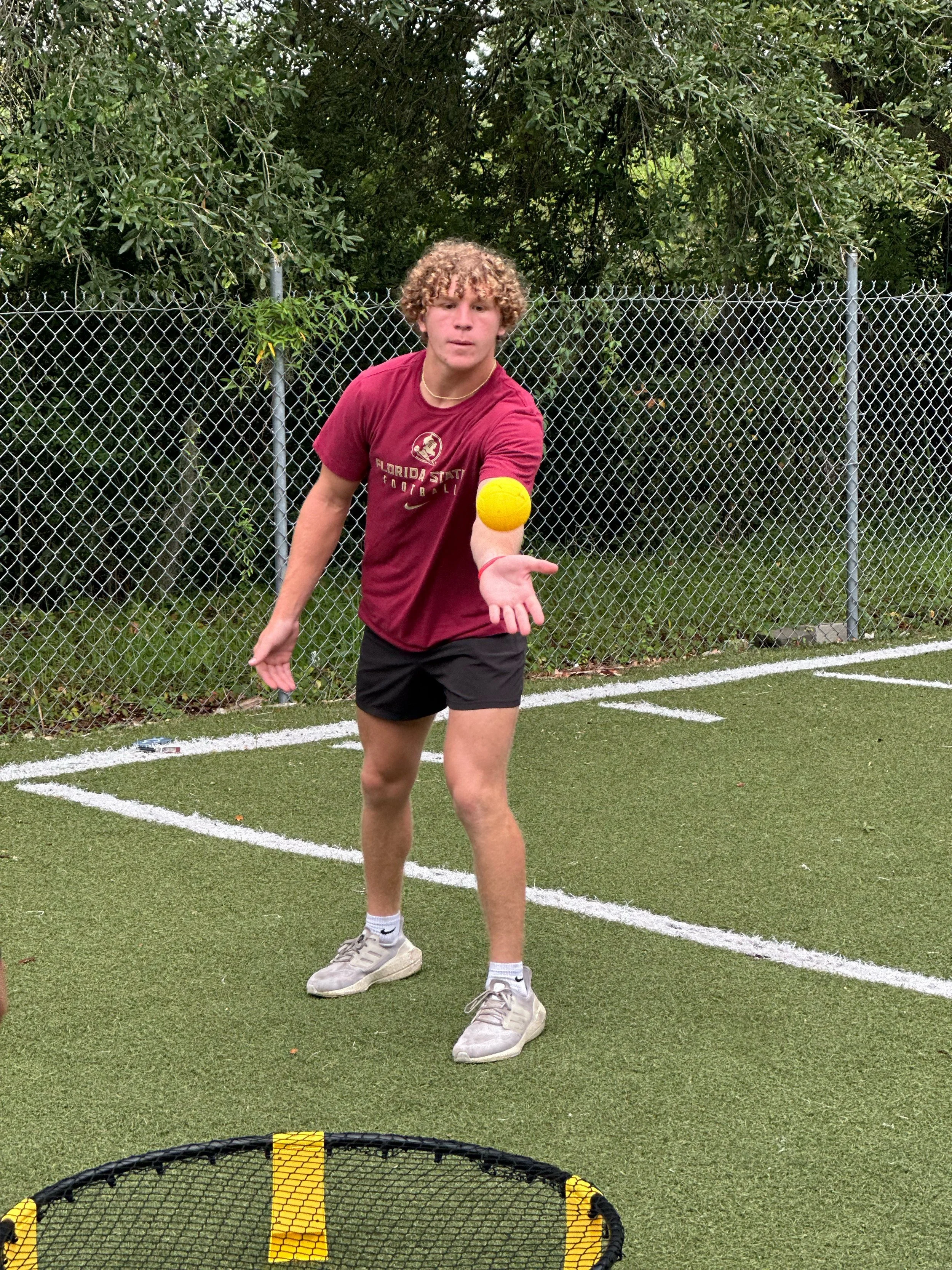 A young man with curly blonde hair playing pickleball outdoors on a court with a chain-link fence and green trees in the background. He is wearing a maroon Florida State t-shirt, black shorts, white socks, and gray sneakers. He is preparing to hit a 