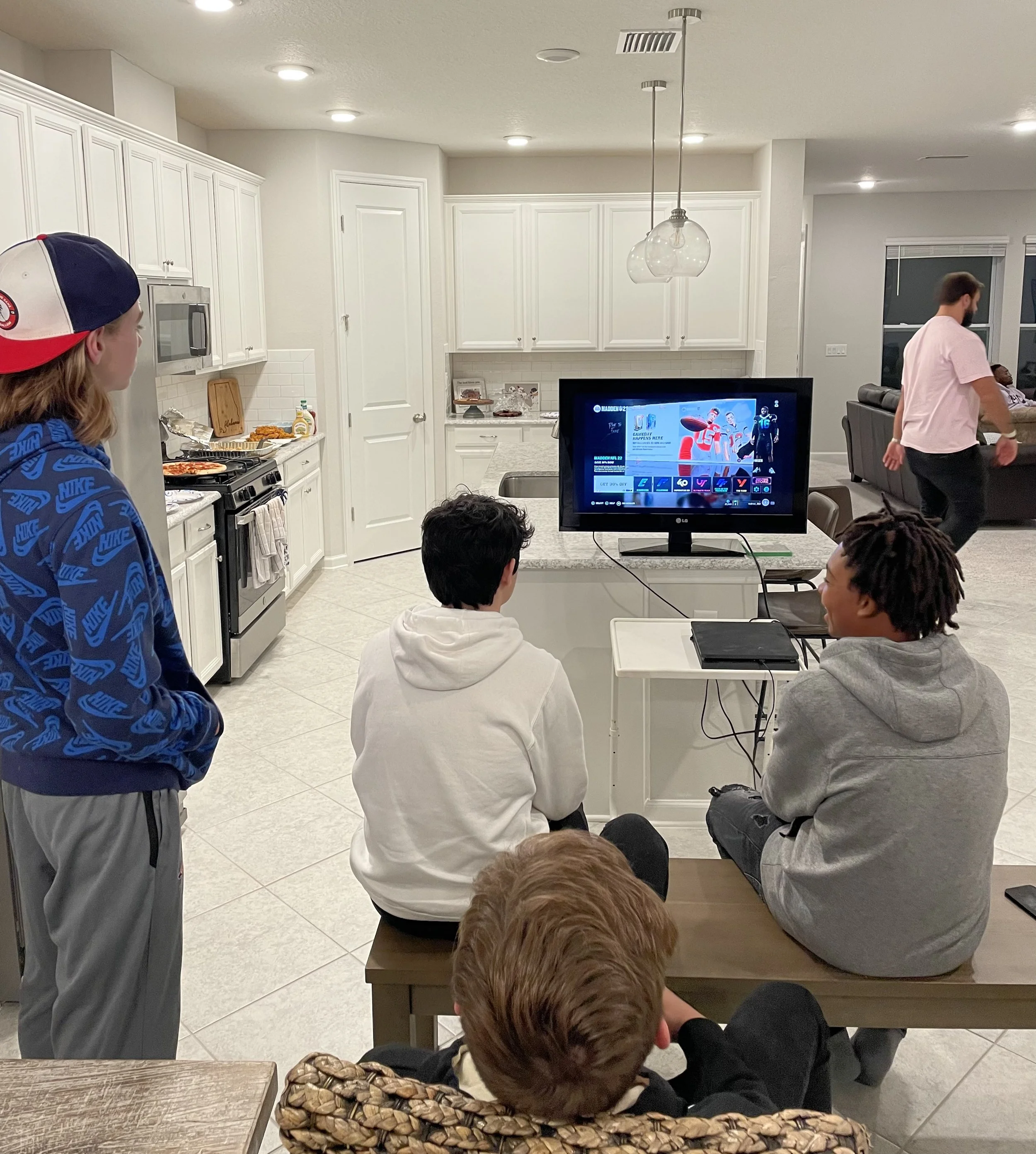 Group of people gathered in a modern kitchen watching TV, with some seated on a bench and others standing.