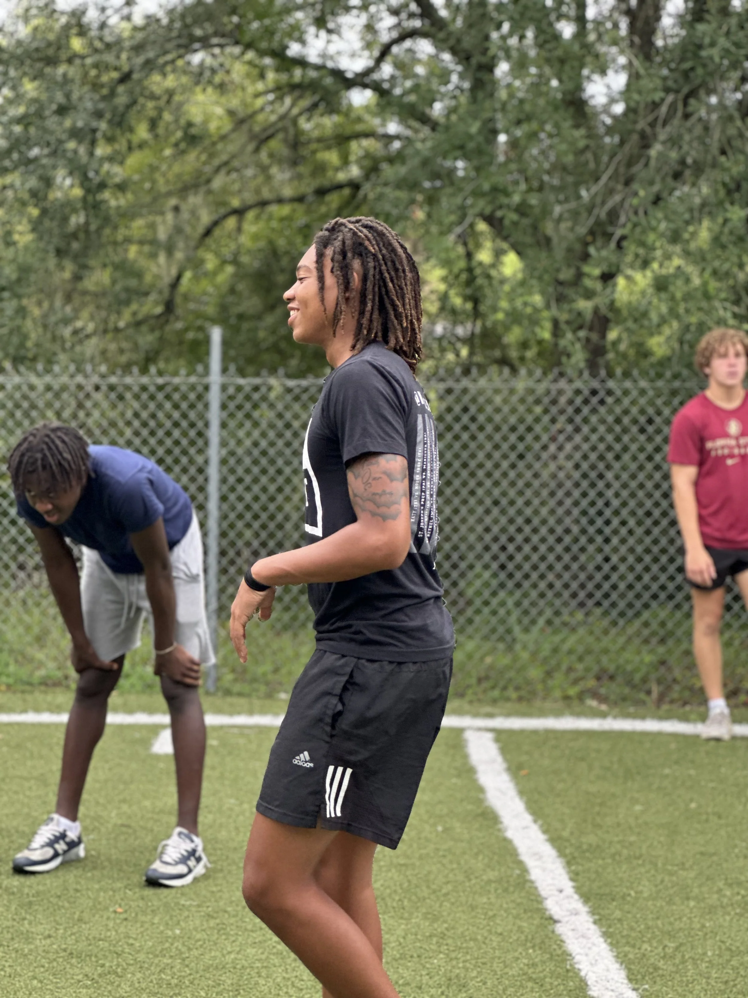 A woman with dreadlocks and tattoos on her arm, wearing a black athletic shirt and shorts, stands on a grassy sports field with a chain-link fence and trees in the background, smiling and possibly talking or laughing. Two other young men are in the b