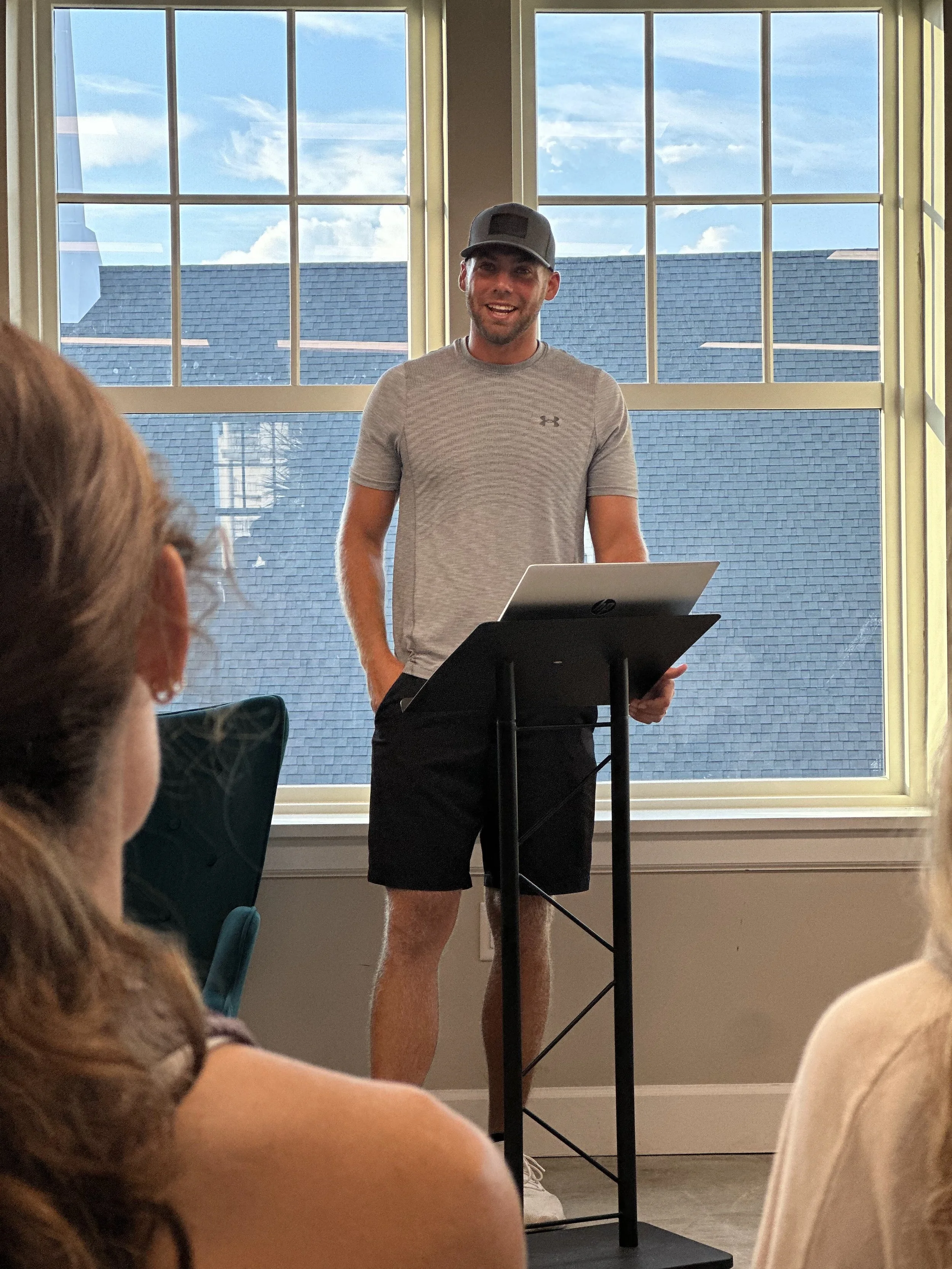 A young man standing behind a lectern with an HP laptop, speaking to an audience in a room with a large window showing a blue sky with clouds and rooftops.