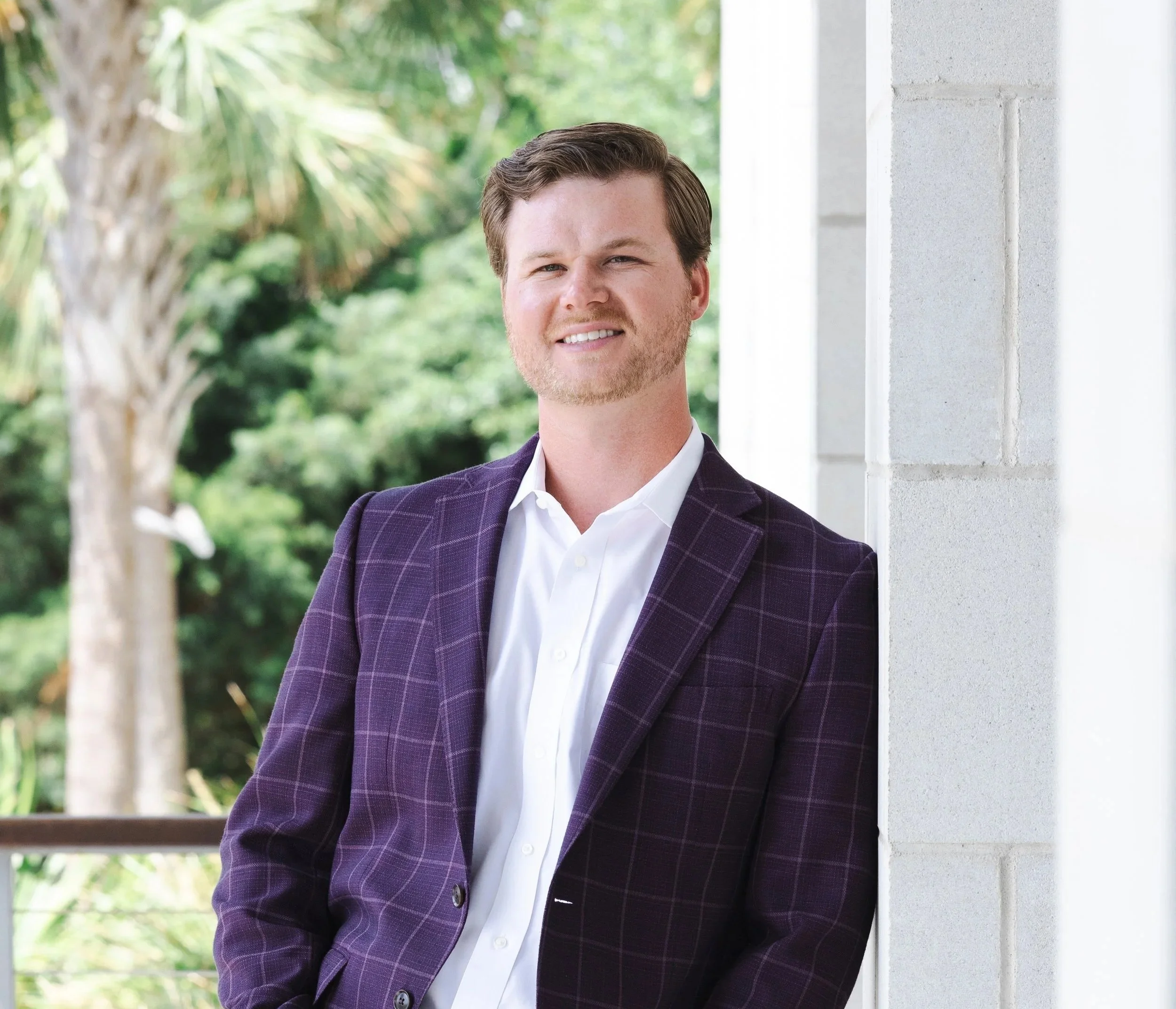 A man with light skin and short brown hair, wearing a white dress shirt and a purple plaid blazer, smiling while leaning against a white concrete wall outdoors surrounded by greenery.