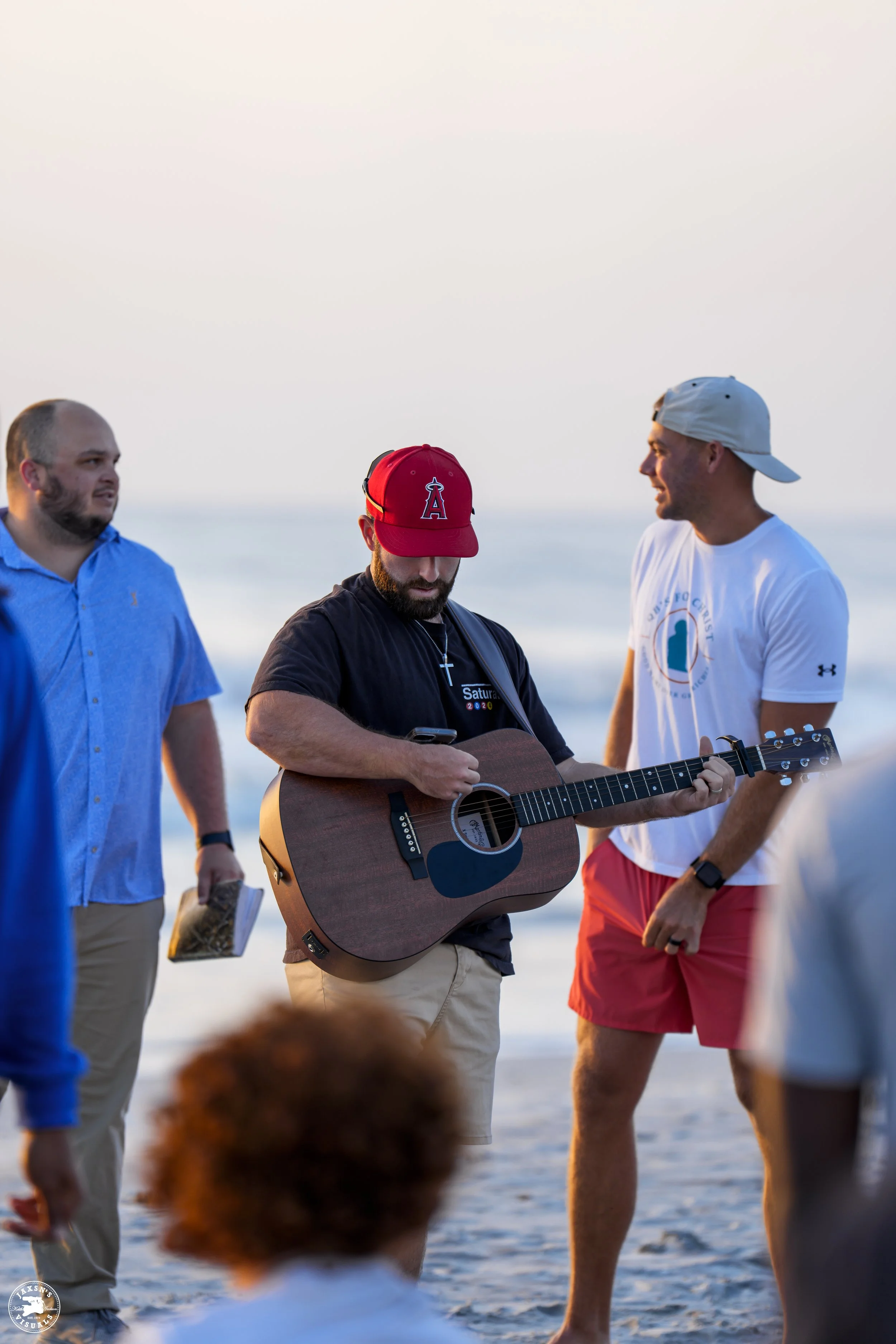 Group of men standing on a beach, one playing guitar, during a sunset or sunrise.