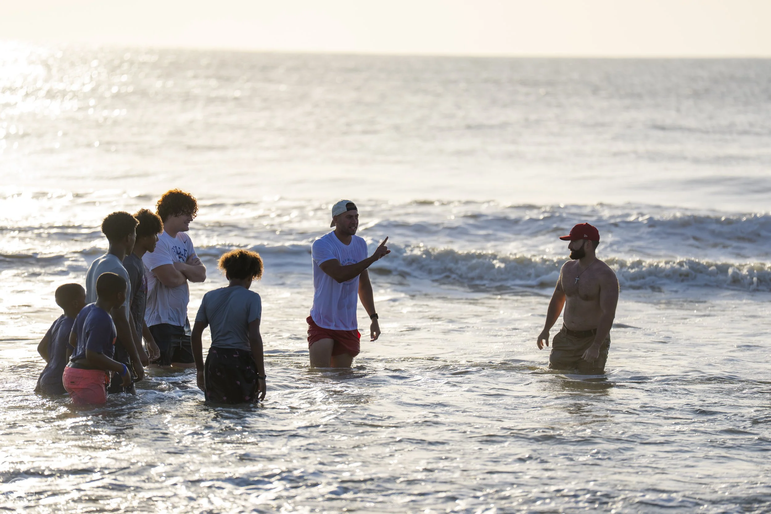 Group of people at the beach, with two men talking in the water while a boy listens, during sunset.