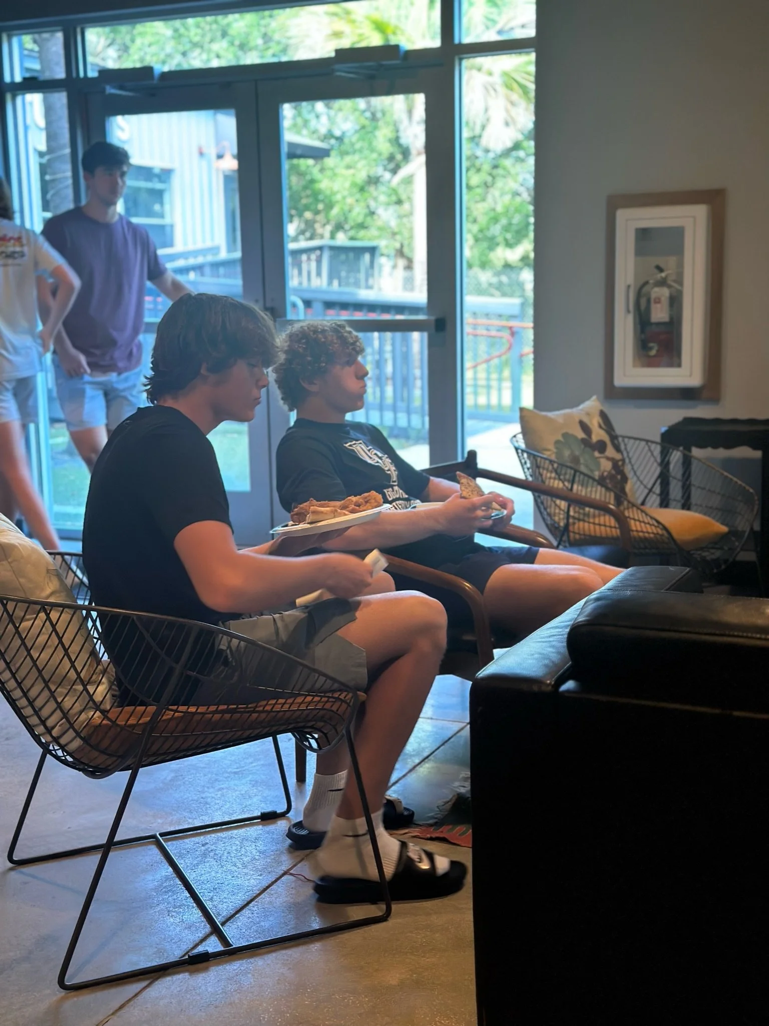 Two young men sitting on chairs inside a cafe, one holding a plate of food, with large glass doors and greenery outside.