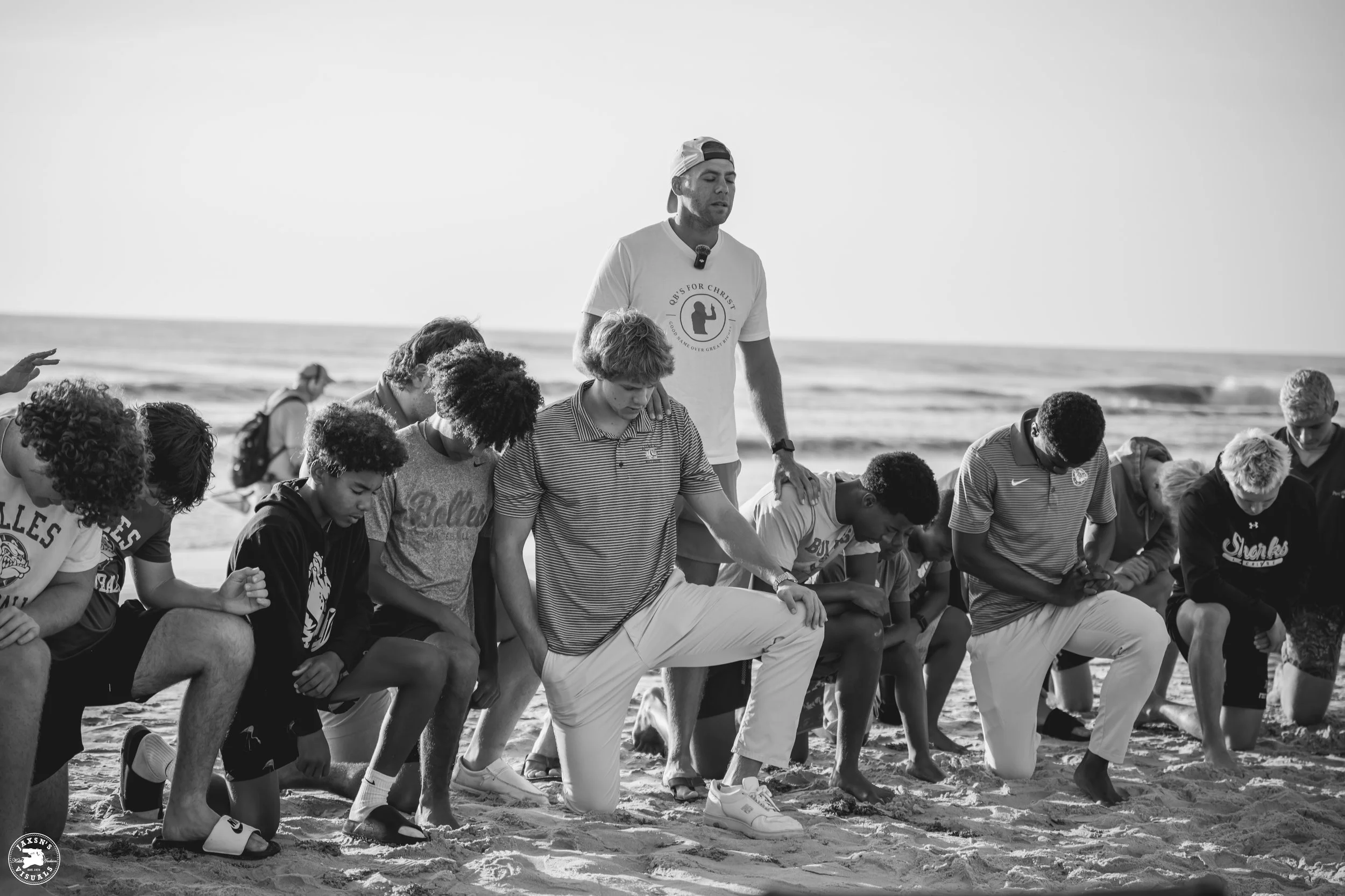 A group of people kneeling on the sand at the beach during a prayer or moment of reflection, with the ocean in the background.