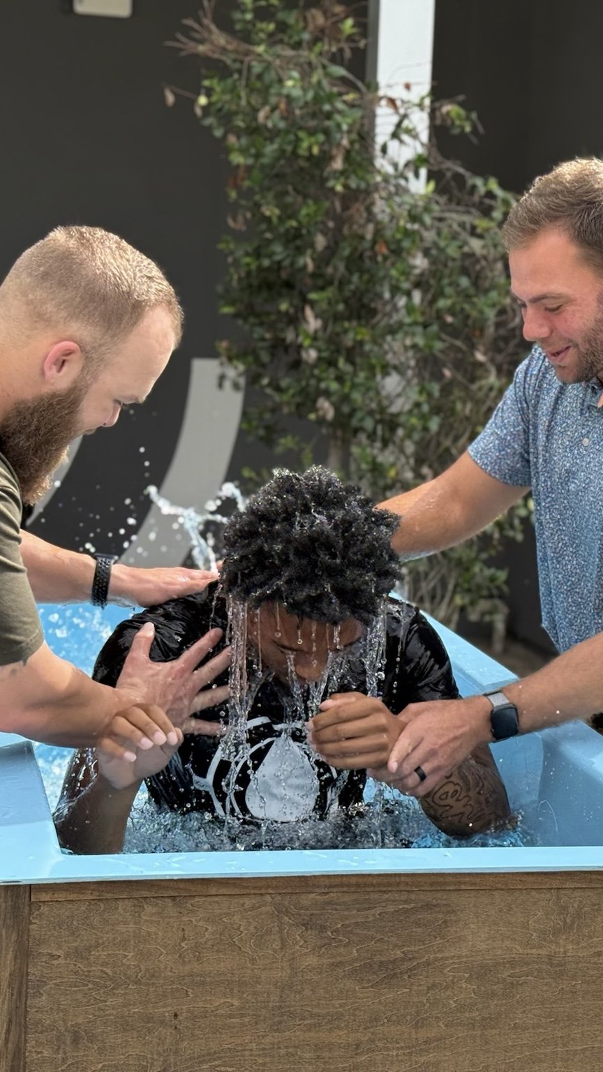 A person with curly hair is being baptized in an indoor water basin while two men assist and support them.